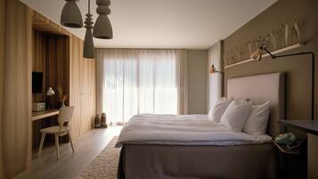 A modern bedroom with a large bed, a desk, and a chair. There is a window with curtains and a wall-mounted shelf with decorative items.