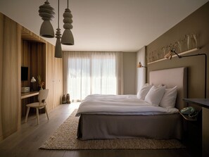 Un dormitorio moderno con una cama grande, un escritorio y una silla. Hay una ventana con cortinas y un estante en la pared con objetos decorativos.