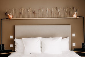 Una cama con ropa blanca y almohadas, un cabecero color beige y un estante de pared con flores secas.