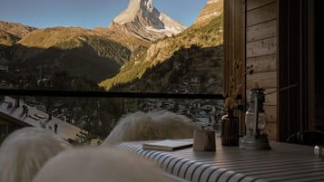 A room with a view of a mountain peak, a wooden interior, and a table with a book and a lamp.