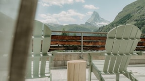 A terrace with two white Adirondack chairs, a wooden table, and a view of a mountain range. - OVERLOOK Lodge by CERVO Zermatt (Zermatt)