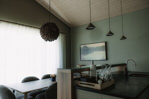 Una cocina moderna con comedor, techo de madera, lámparas colgantes, un ventanal con cortinas translúcidas y un cuadro de paisaje en la pared.
