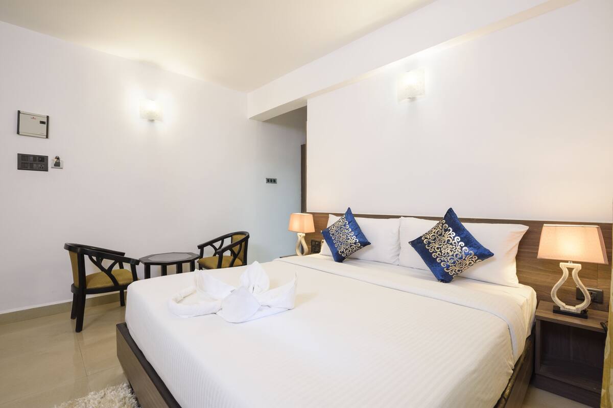 A hotel room with a neatly made bed, two bedside lamps, a small table with a chair, and a wall-mounted light fixture.