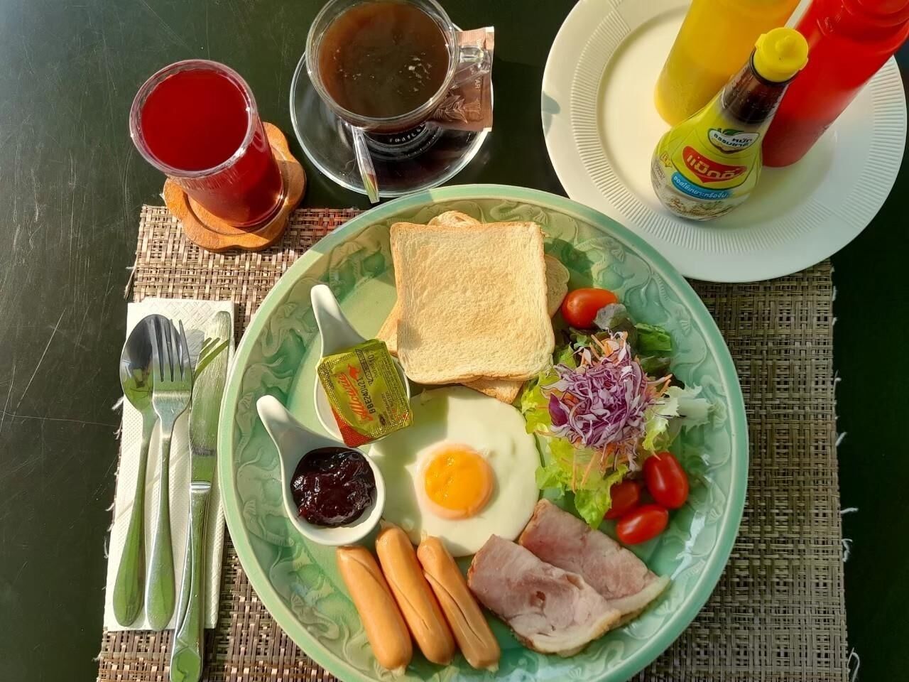 Un plato con tostadas, huevo, jamón, salchichas y ensalada, junto con salsas y bebidas sobre una mesa.