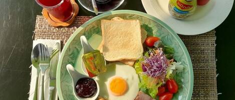 Un plato con tostadas, huevo, jamón, salchichas y ensalada, junto con salsas y bebidas sobre una mesa.
