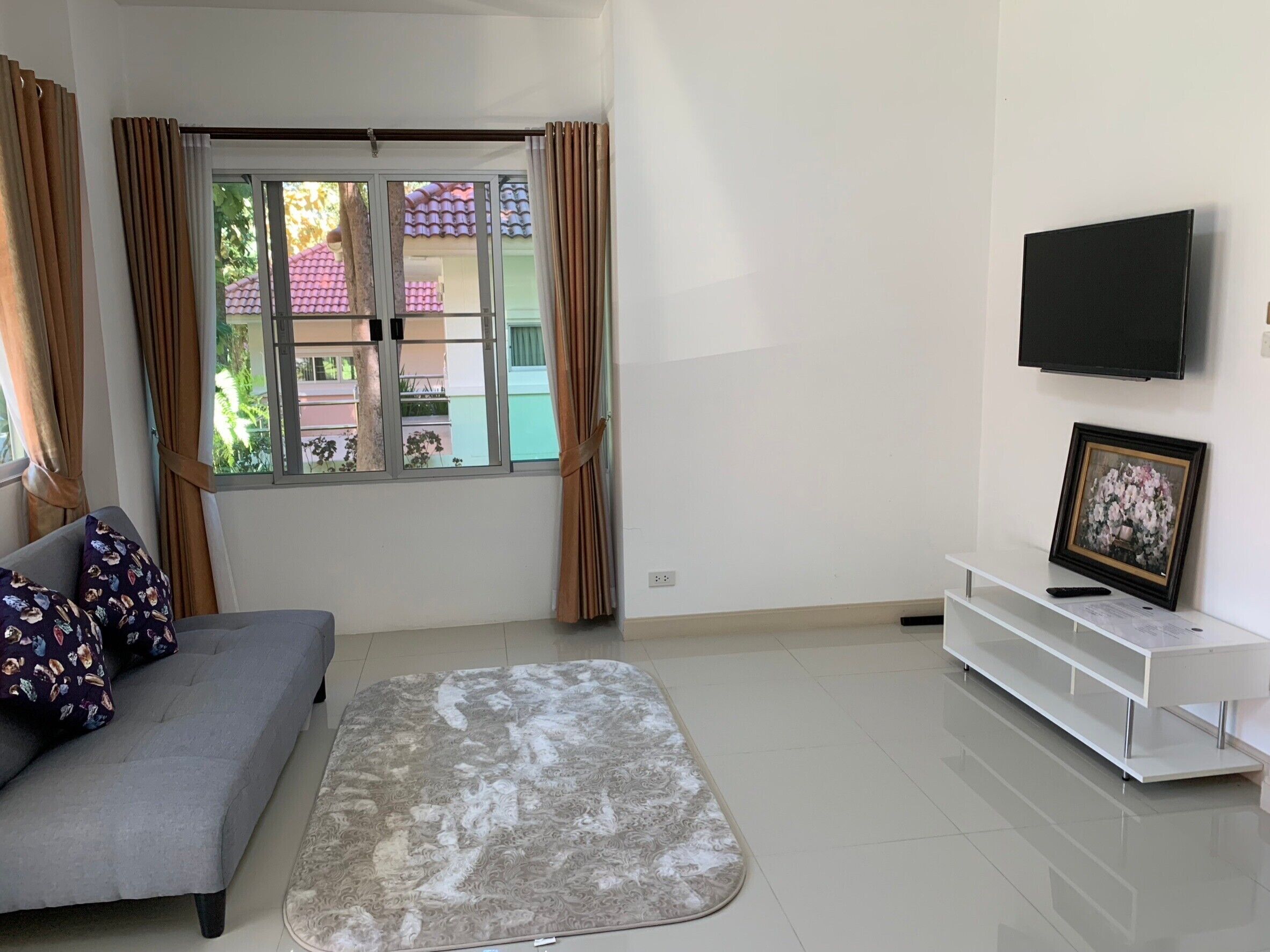 A living room with a grey sofa, patterned cushions, a grey rug, a TV on the wall, and a framed picture on a white shelf.
