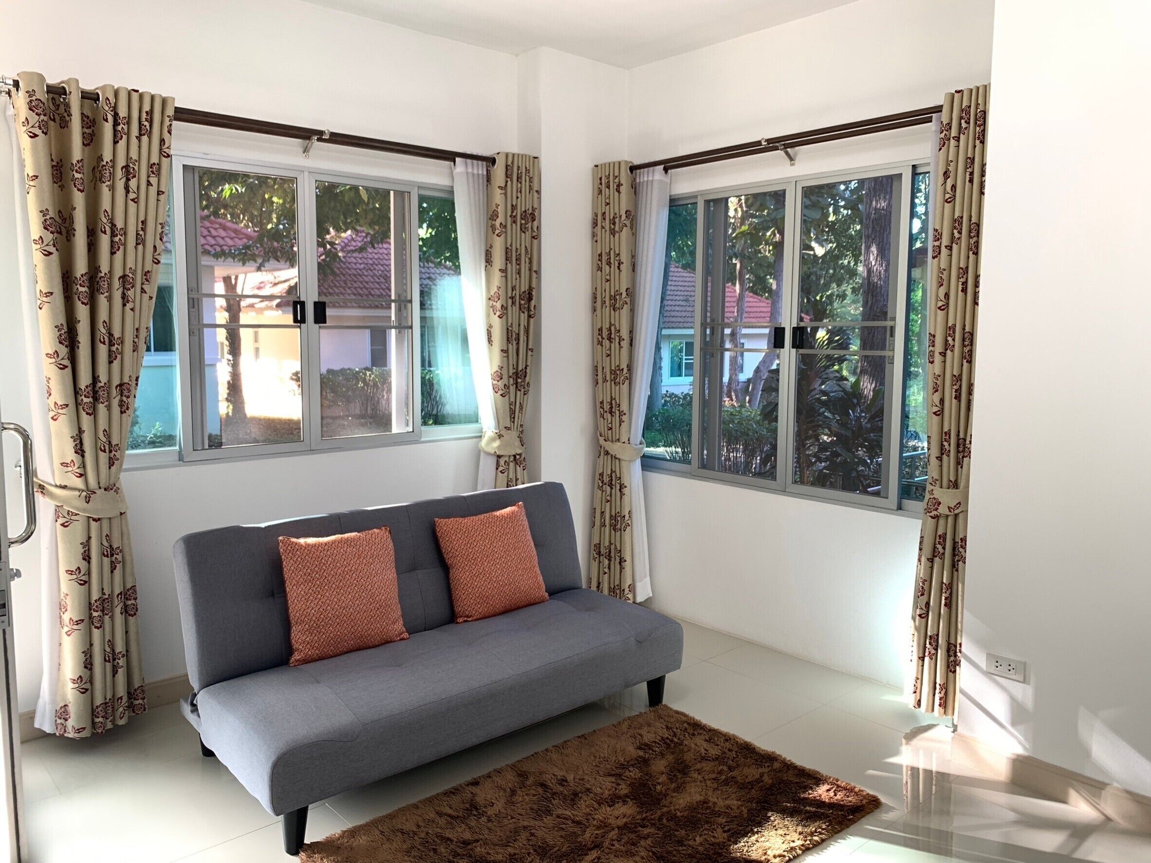 A room with a grey sofa, two patterned curtains, and a view of greenery outside.