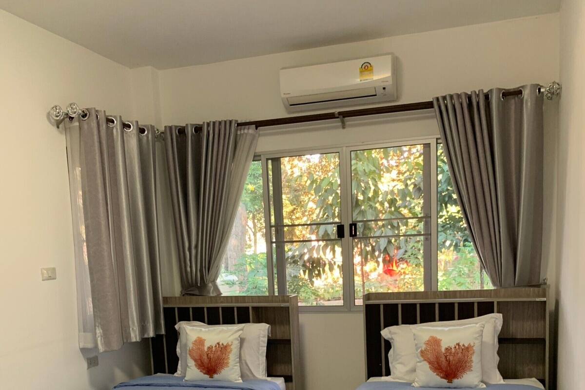 A room with two single beds, grey curtains, a window with a view of greenery, and an air conditioning unit.