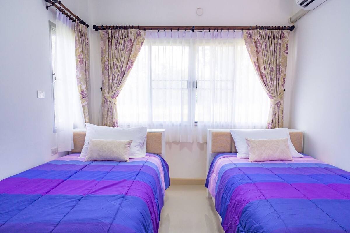 A room with two beds, each with a purple and blue bedspread, white pillows, and a wooden headboard. There are two windows with floral curtains and a ceiling fan.