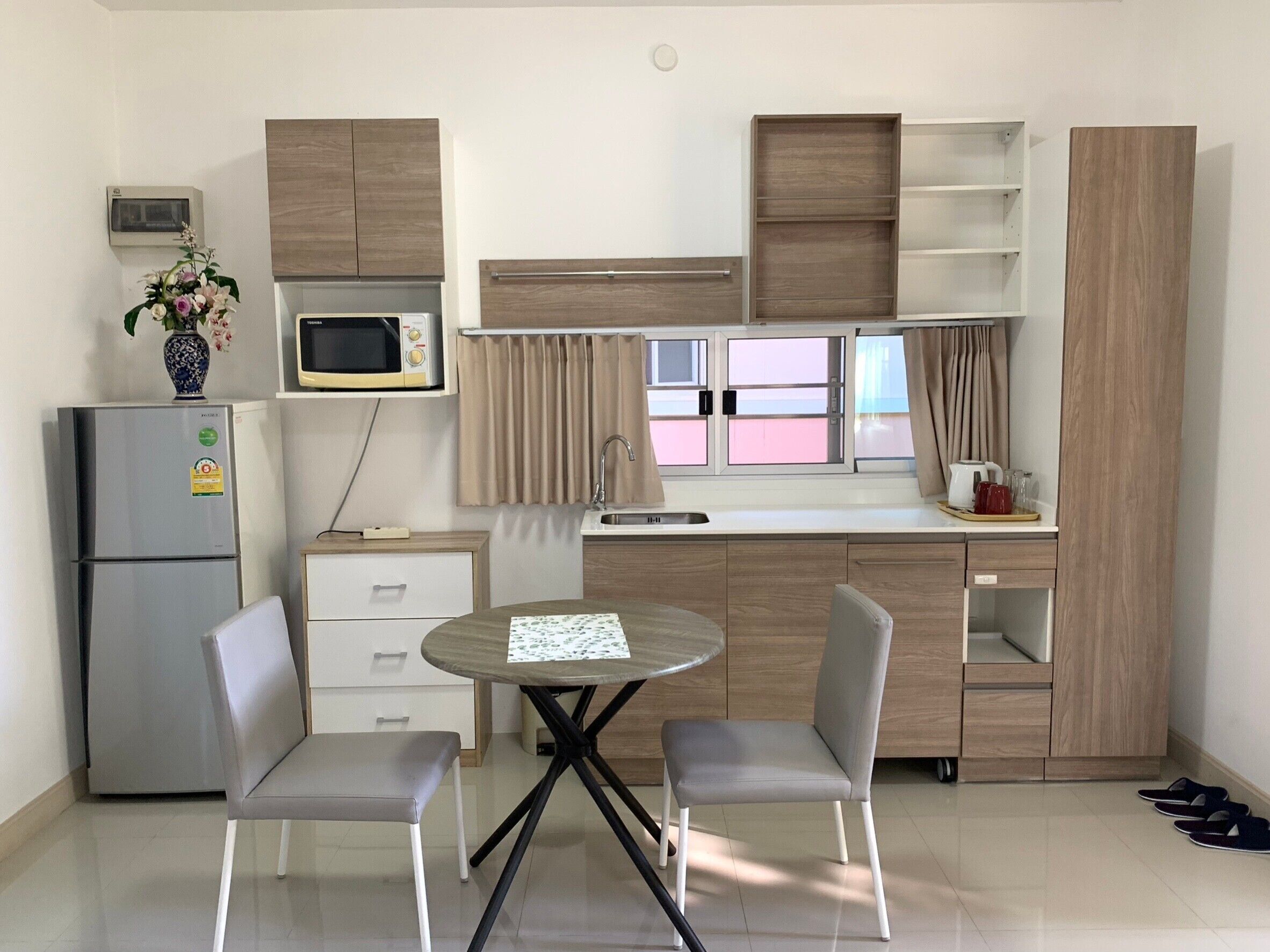 A compact kitchen with a refrigerator, microwave, and wooden cabinets. There is a small dining table with two chairs and a vase with flowers on the counter.