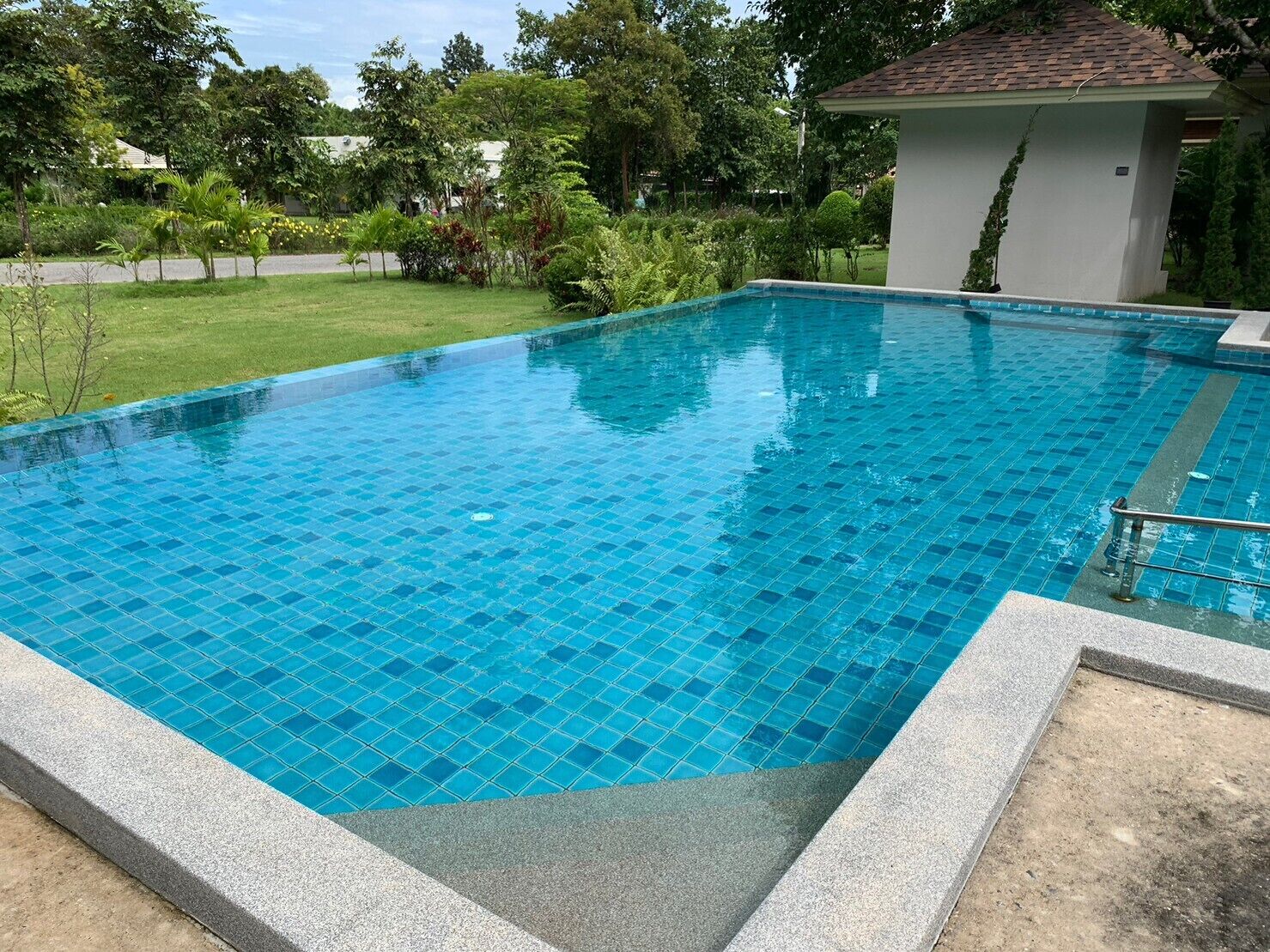 A rectangular swimming pool with blue tiles, surrounded by a concrete deck and a small white building with a tiled roof.