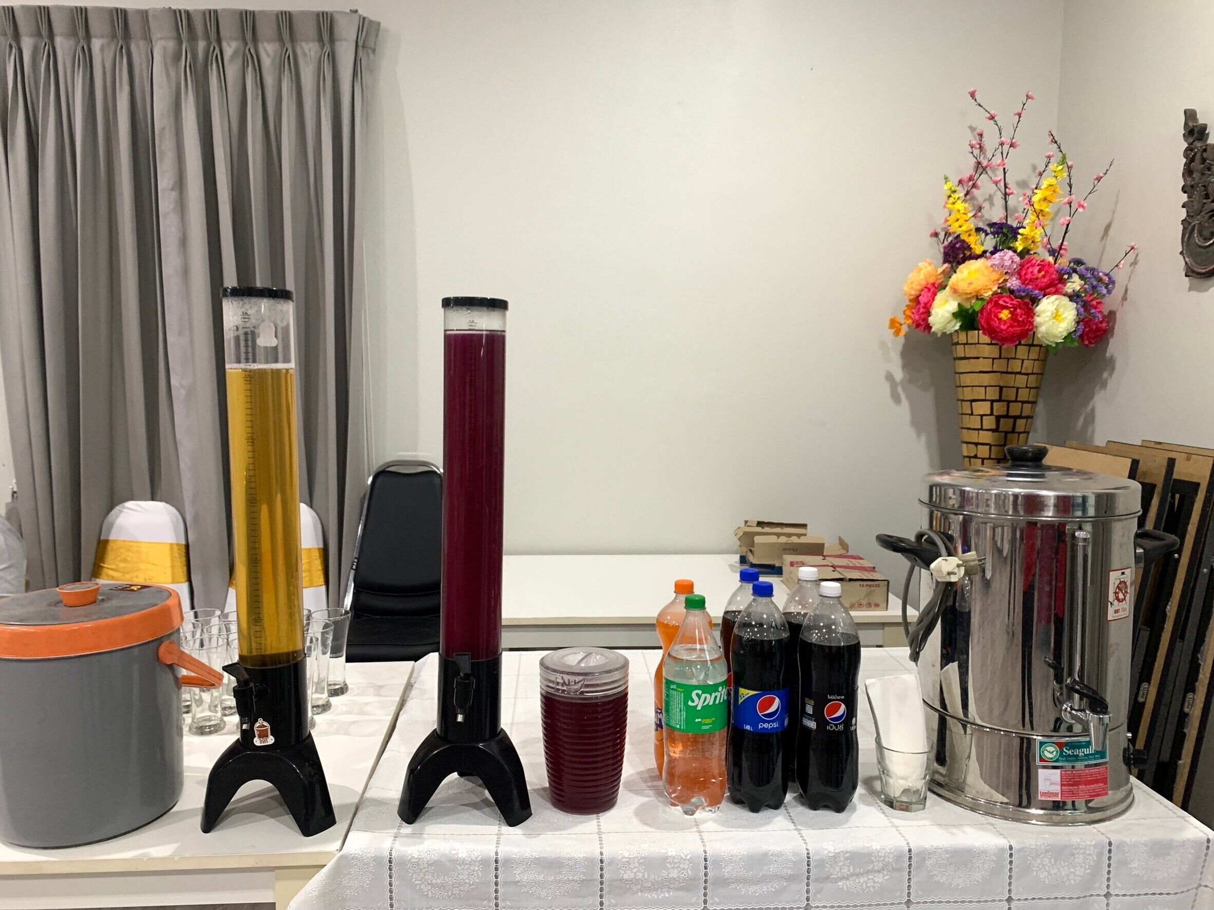 Una mesa con dispensador de agua, un recipiente de vidrio alto y varias botellas de bebidas.