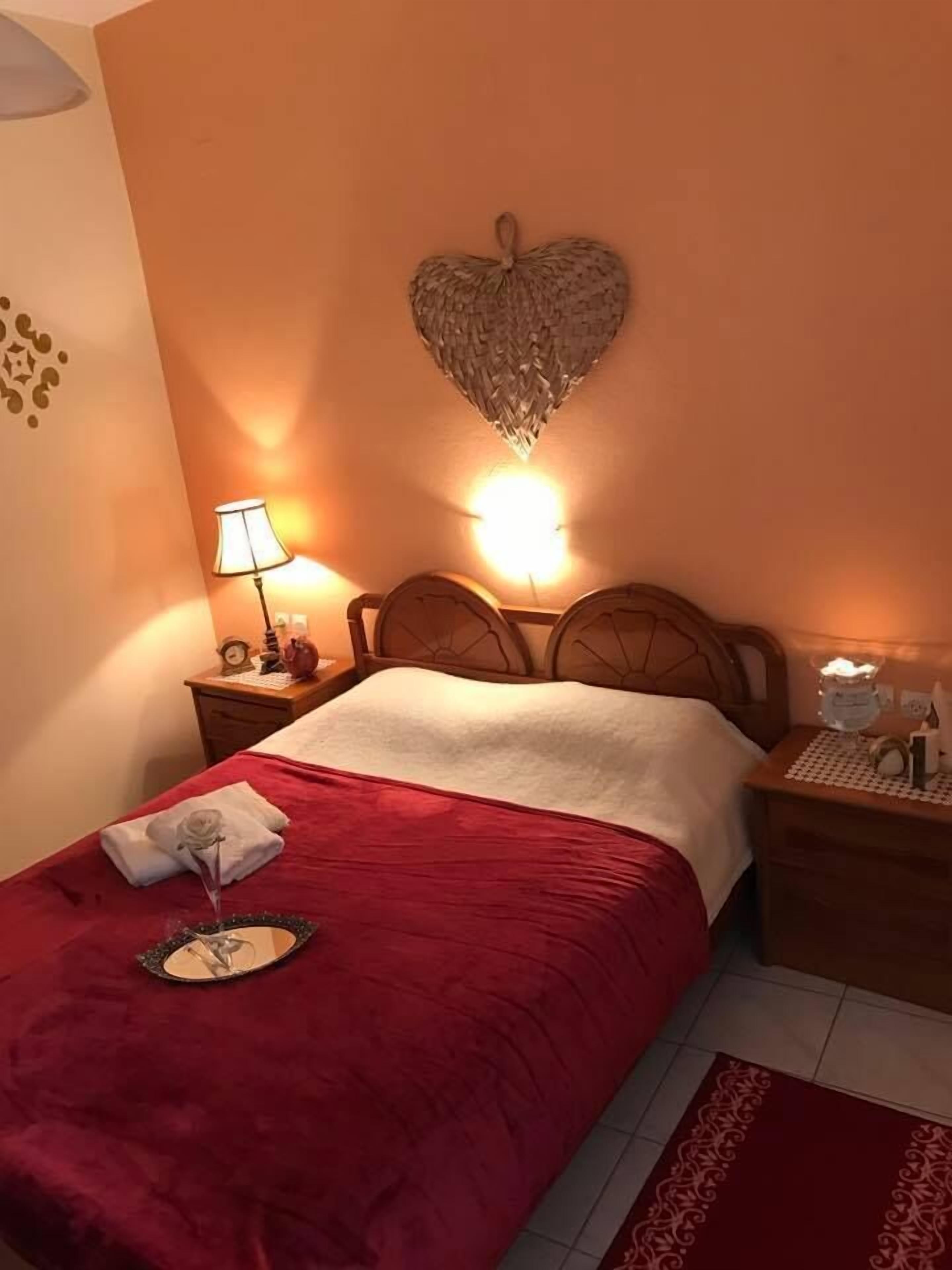 A neatly made bed with a red blanket, a heart-shaped wall decoration, and a bedside table with a lamp and a clock.