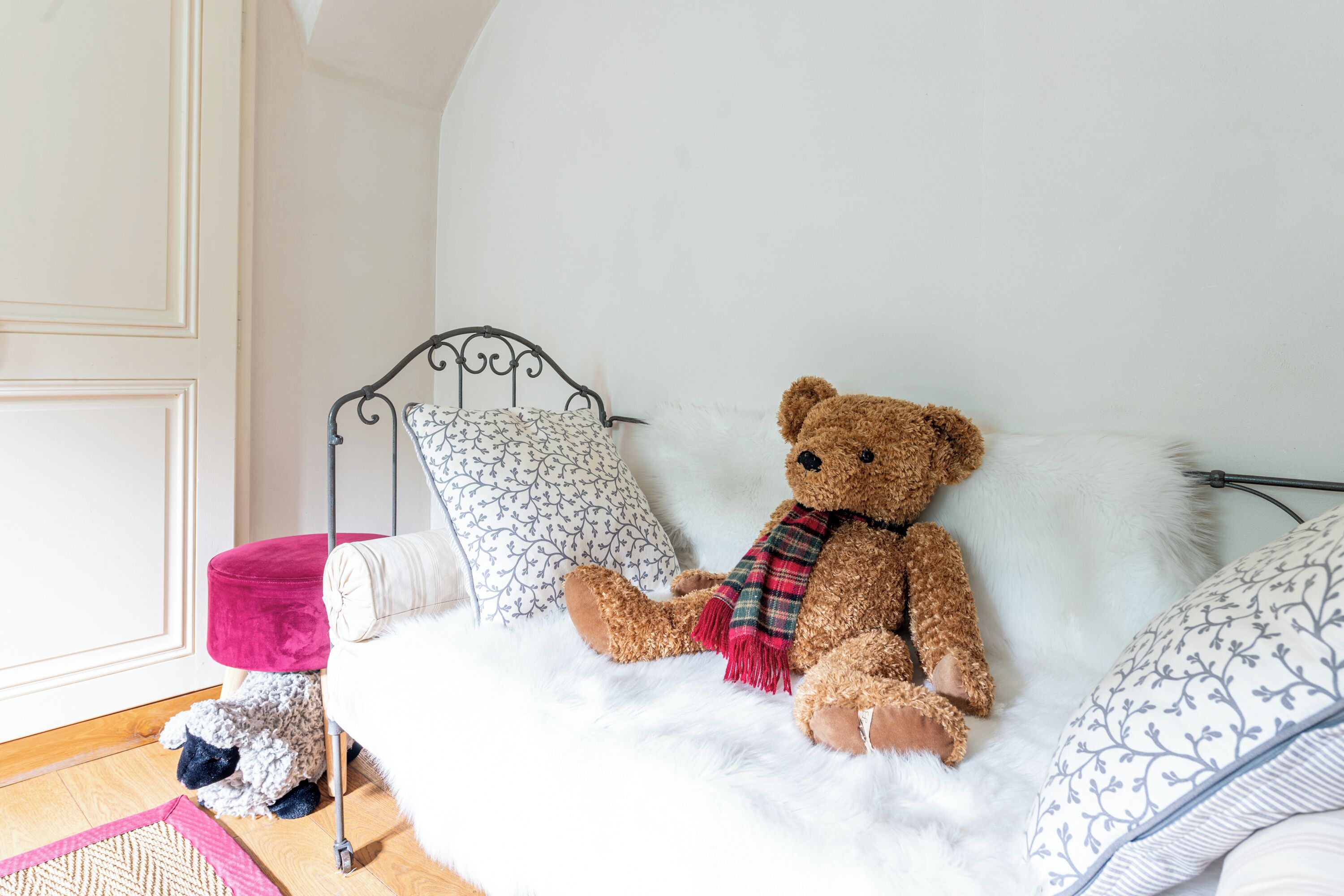 A bed with a teddy bear, pillows, and a small table with a pink cushion.
