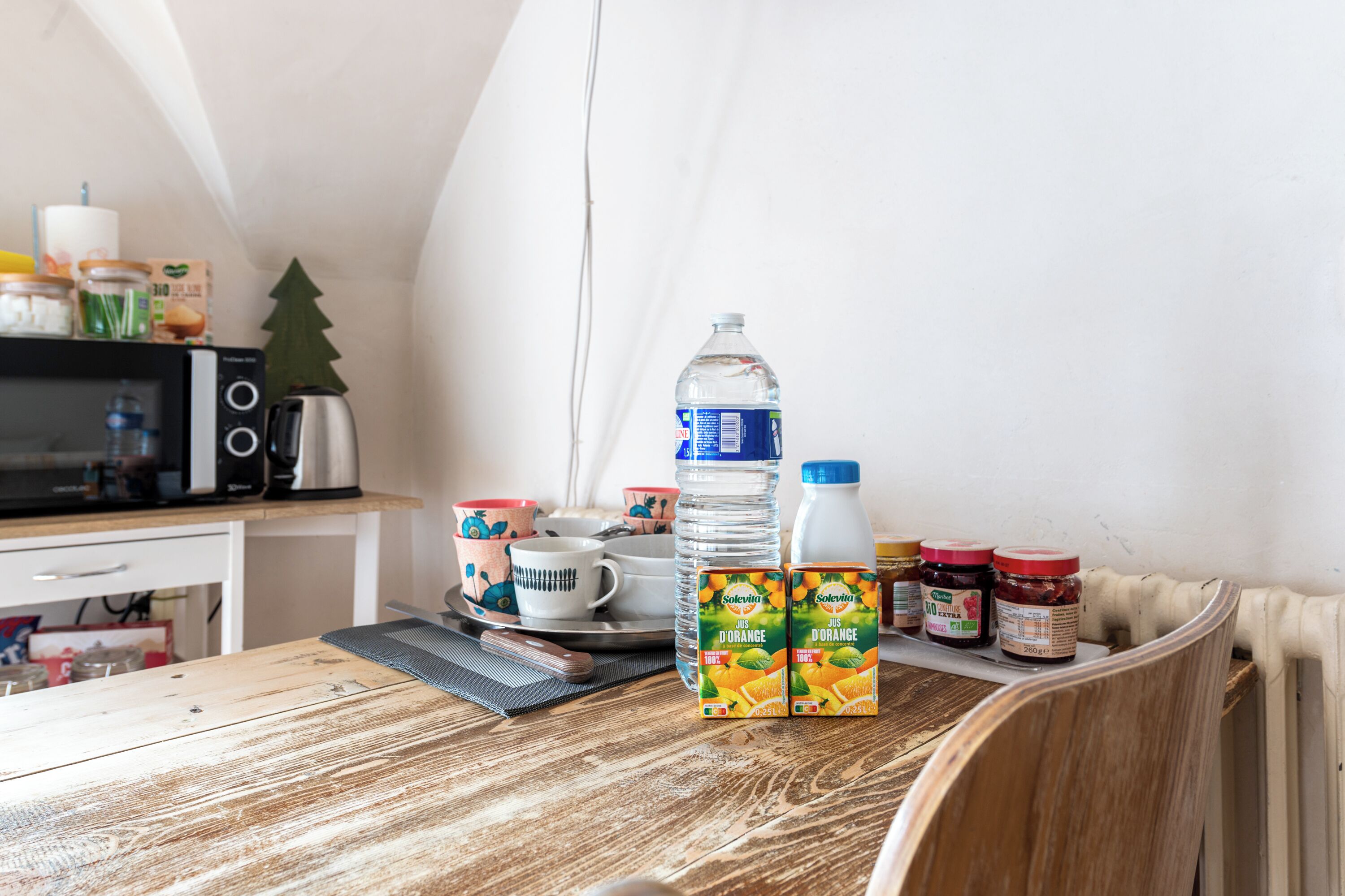 A kitchen with a wooden table, a microwave, a kettle, and various food items including juice boxes and jars.