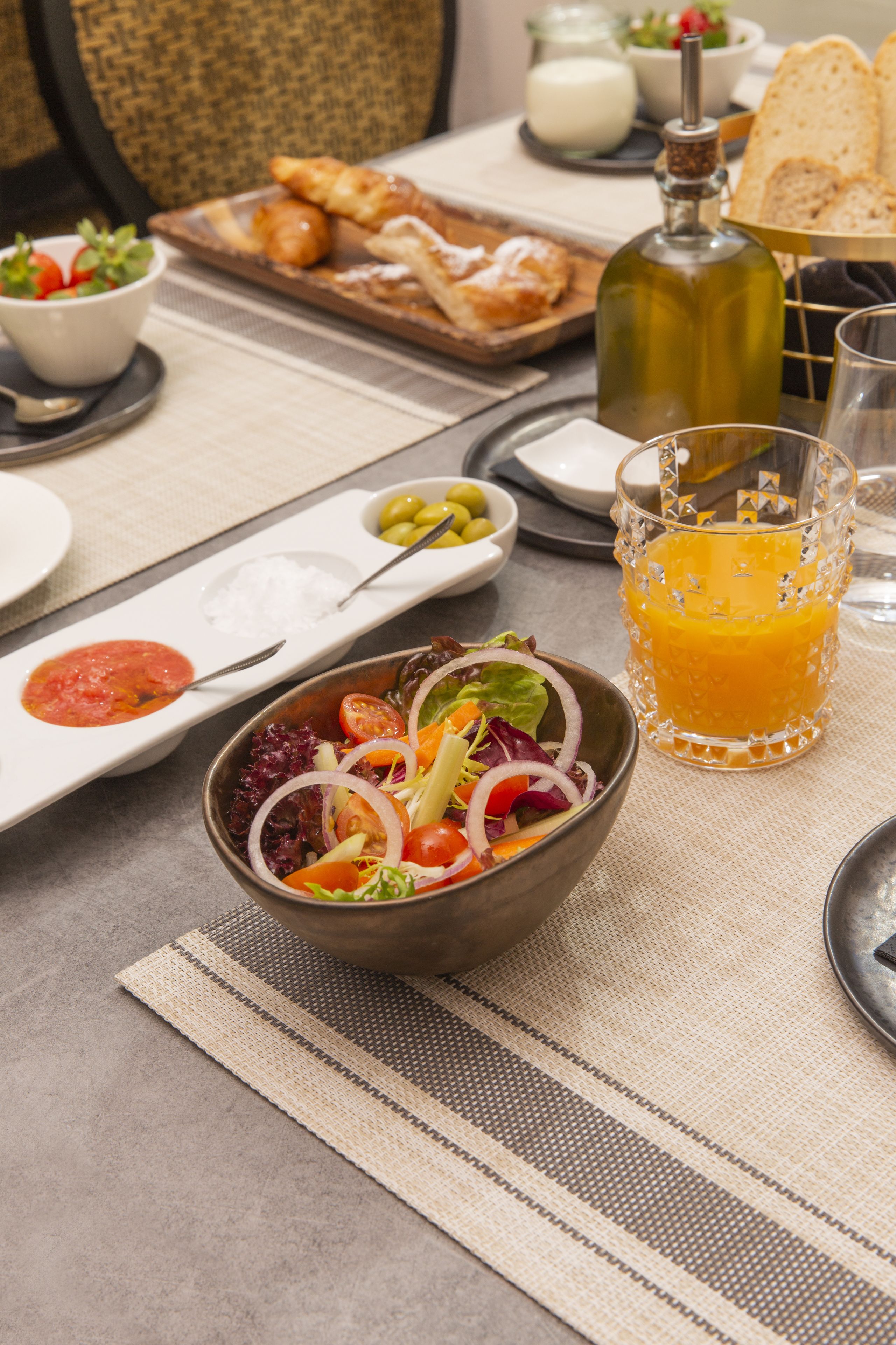Une table à manger avec un saladier rempli de salade, un verre de jus d’orange et une bouteille d’huile d’olive.