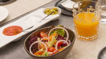 Une table à manger avec un saladier rempli de salade, un verre de jus d’orange et une bouteille d’huile d’olive.