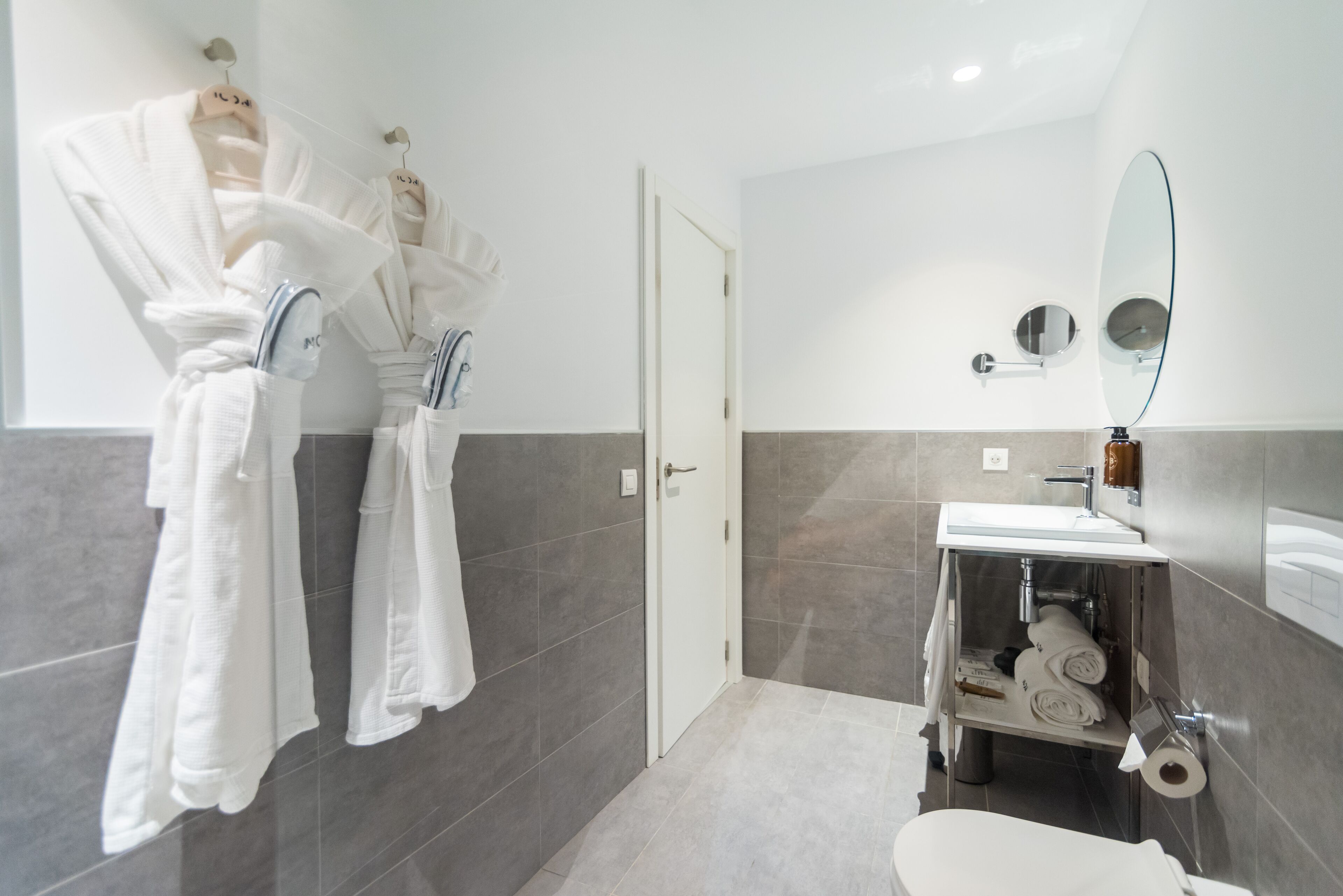 Un baño moderno con un lavamanos blanco, un espejo y un inodoro. Hay toallas colgadas en ganchos y una repisa con artículos de tocador.