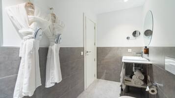 Un baño moderno con un lavamanos blanco, un espejo y un inodoro. Hay toallas colgadas en ganchos y una repisa con artículos de tocador.