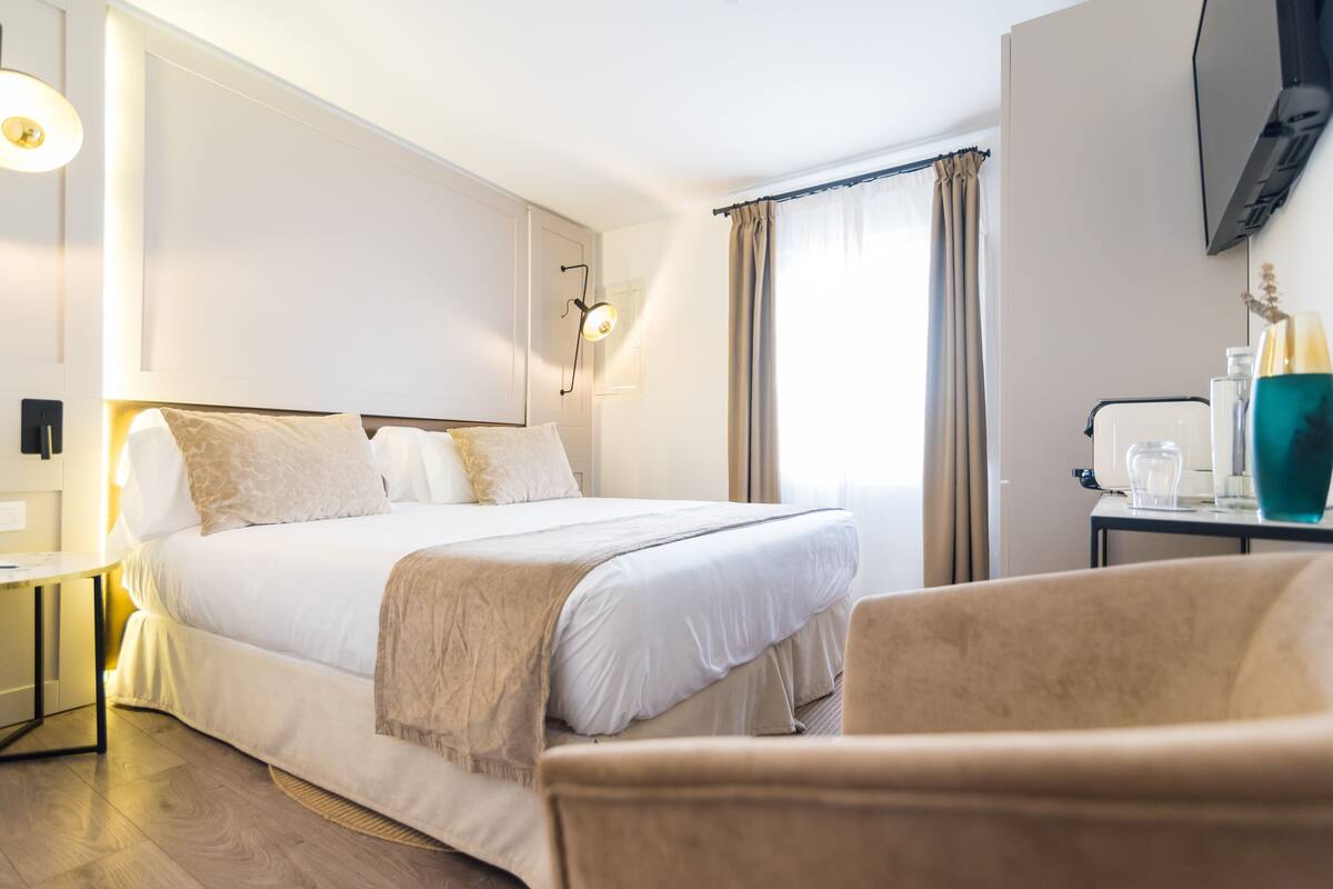 Ein Hotelzimmer mit einem großen Bett, einem Nachttisch, einem Sessel, einem an der Wand montierten Fernseher und einem Fenster mit Vorhängen.
