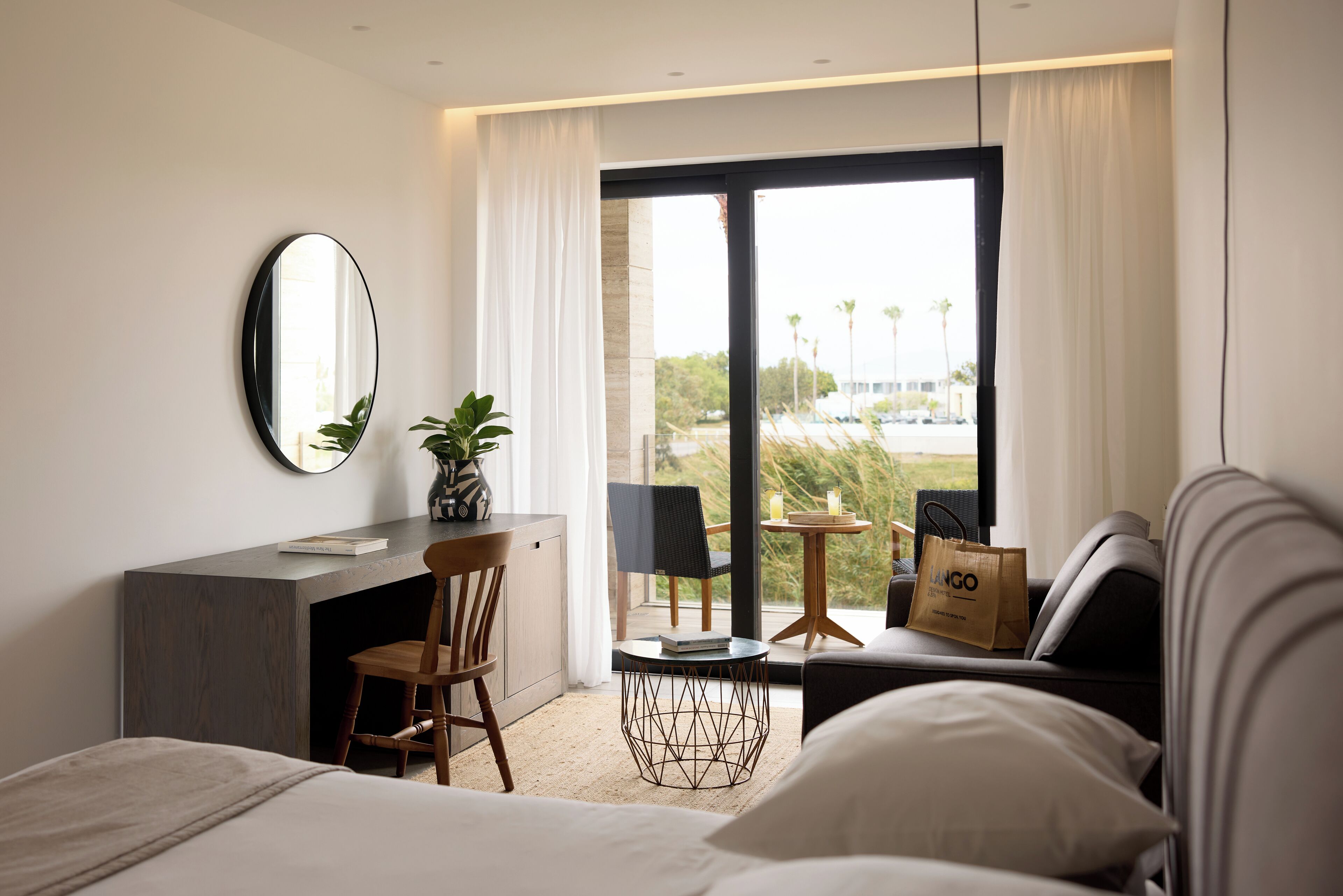 Una habitación de hotel moderna con una cama, un escritorio, una silla, una mesita redonda y un ventanal grande con cortinas.