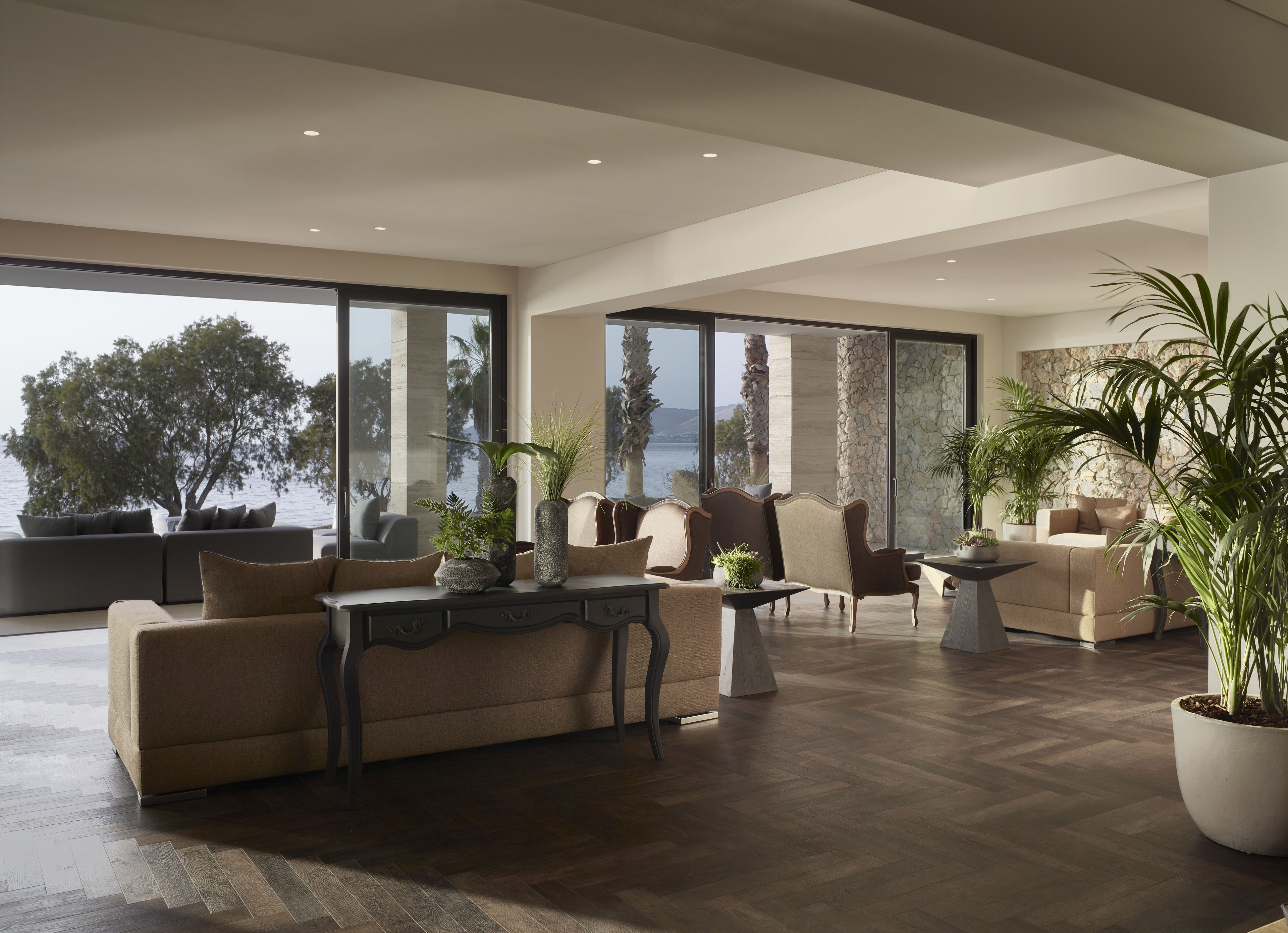 Un salon spacieux avec un grand canapé, des fauteuils et une table basse. On y trouve plusieurs plantes en pot et, grâce aux grandes fenêtres, une vue sur la mer.