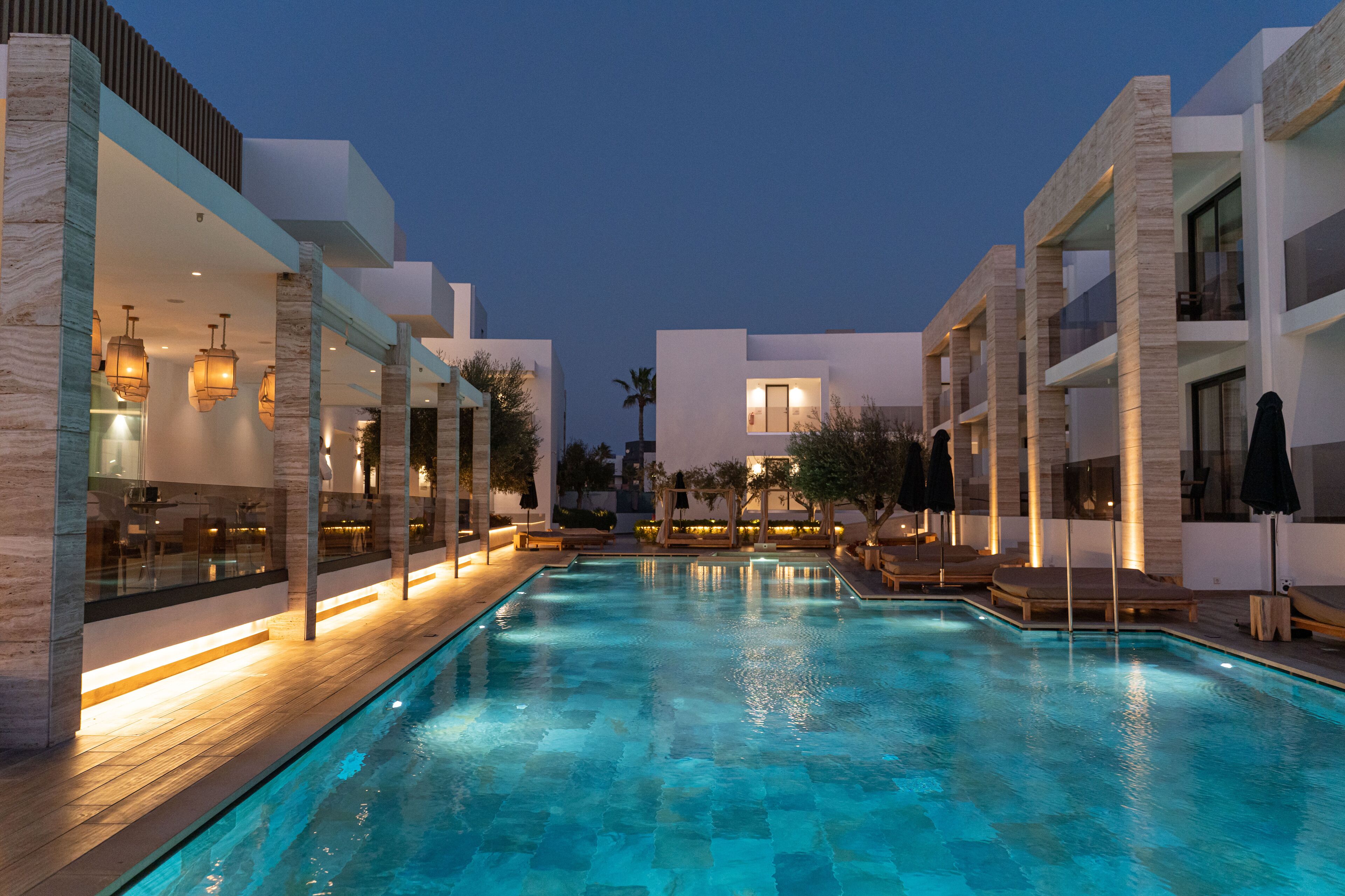 Ein Pool mit Liegestühlen und Sonnenschirmen, umgeben von modernen Gebäuden mit großen Fenstern und Außenbeleuchtung.