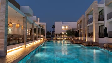 Ein Pool mit Liegestühlen und Sonnenschirmen, umgeben von modernen Gebäuden mit großen Fenstern und Außenbeleuchtung.