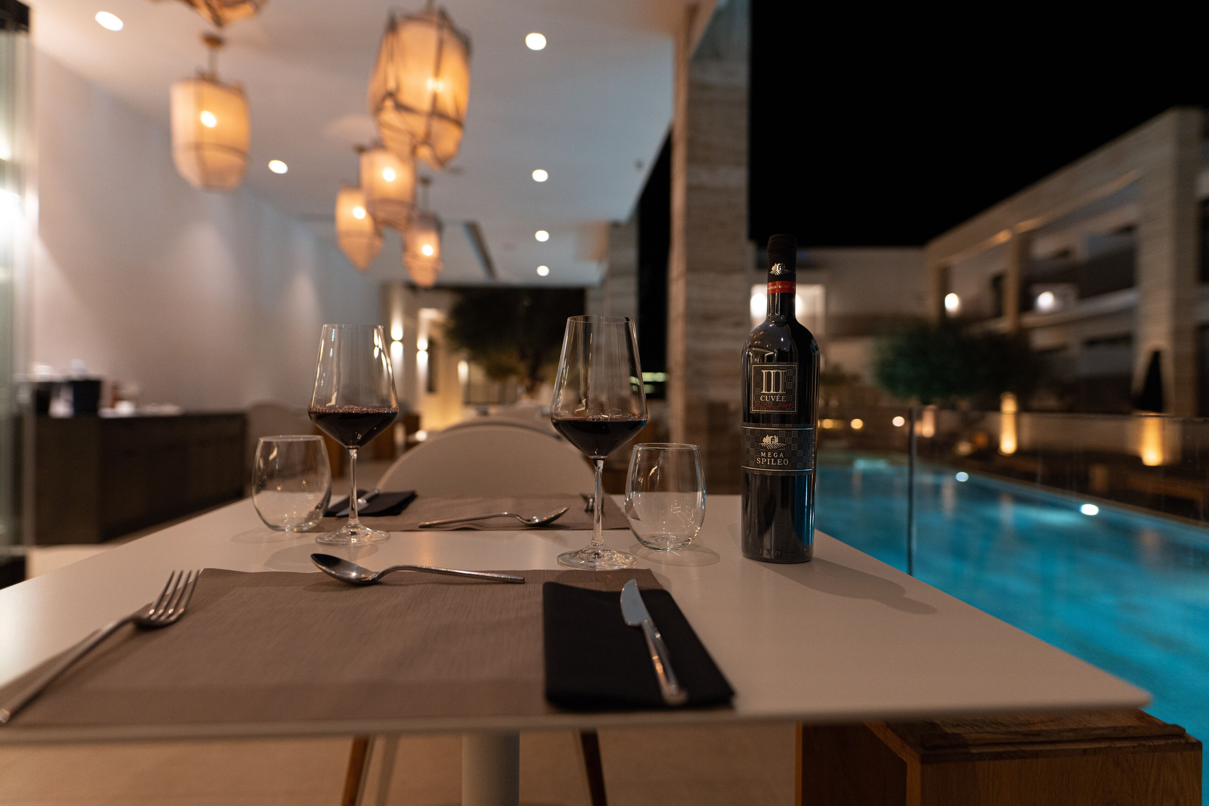 A dining table set with wine glasses, a bottle of wine, and cutlery, overlooking a pool.