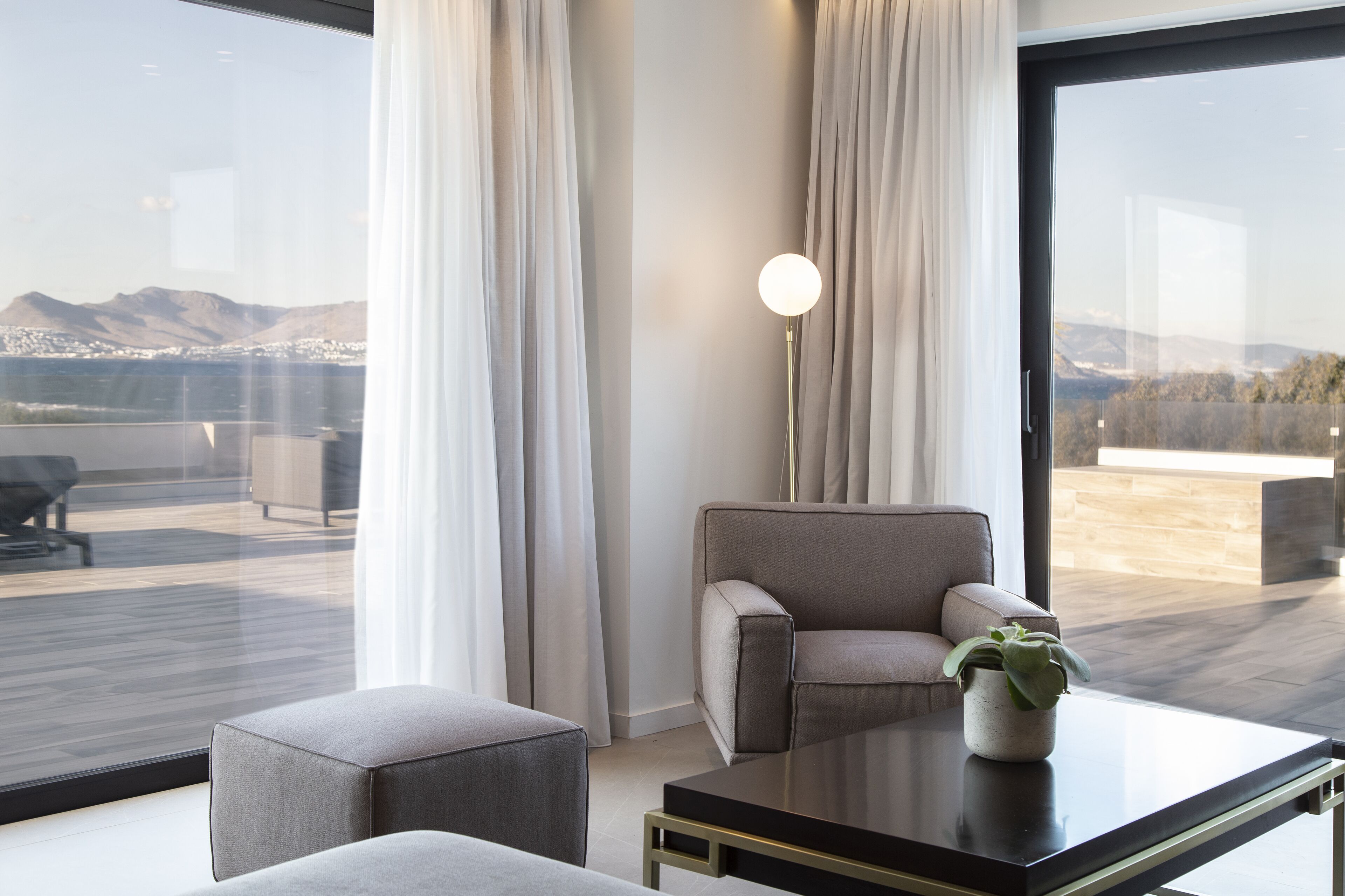 A modern living room with a sofa, a coffee table, and a floor lamp. A large window offers a view of a cityscape and mountains.