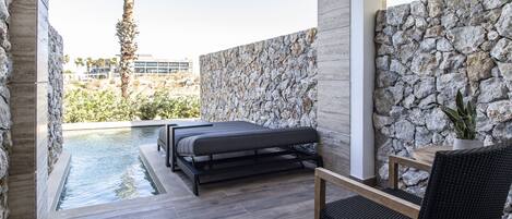 Un área exterior moderna para relajarse, con un muro de piedra, una pequeña piscina, una cama y una mesa de comedor con sillas.