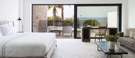 A modern bedroom with a large bed, a dining table, and a sofa. There is a balcony with a view of the sea.