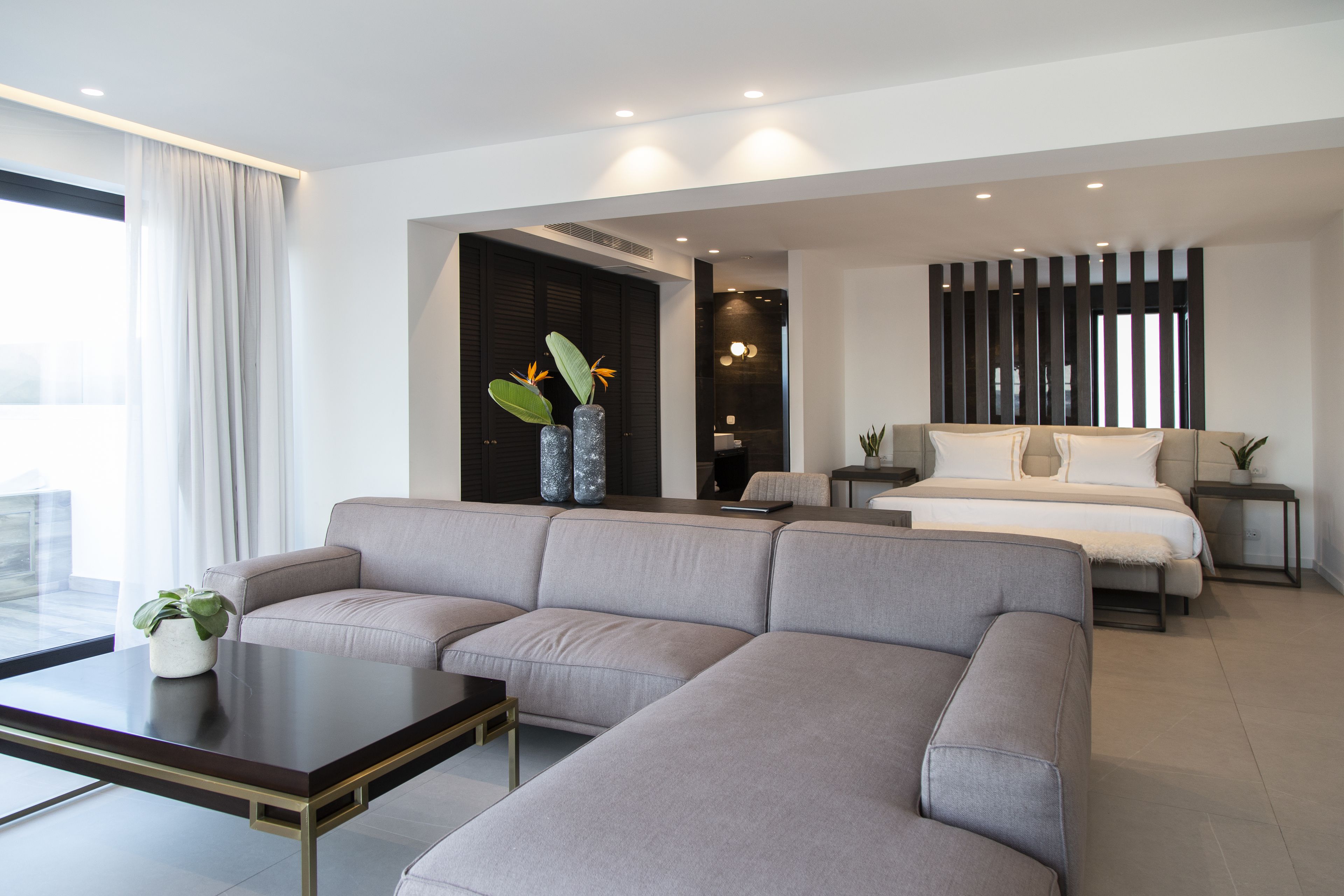 Une chambre d’hôtel moderne avec un canapé d’angle gris, une petite table avec une plante, un lit avec une literie blanche et un paravent sombre.