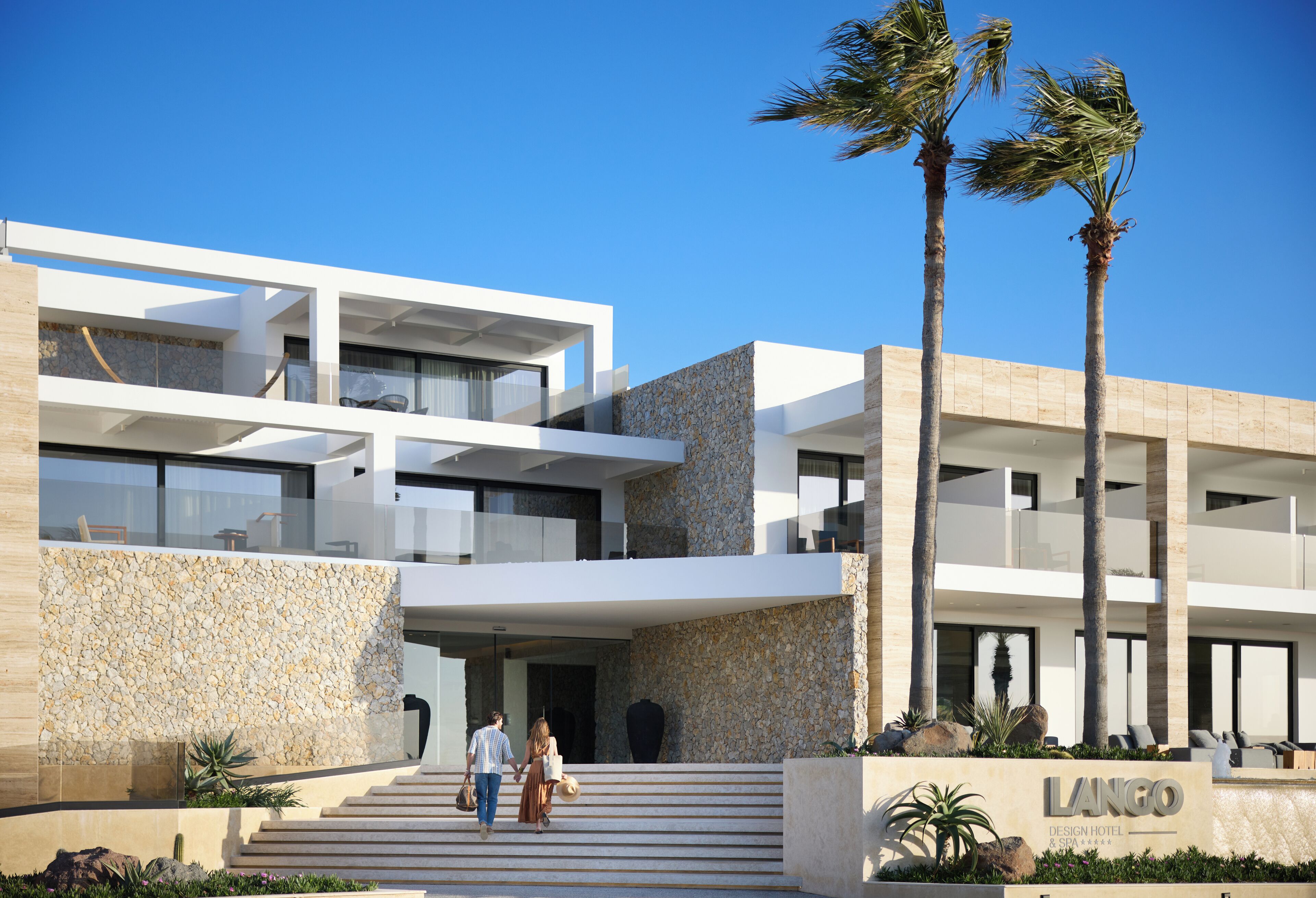 A modern hotel with a stone facade, multiple balconies, and palm trees.