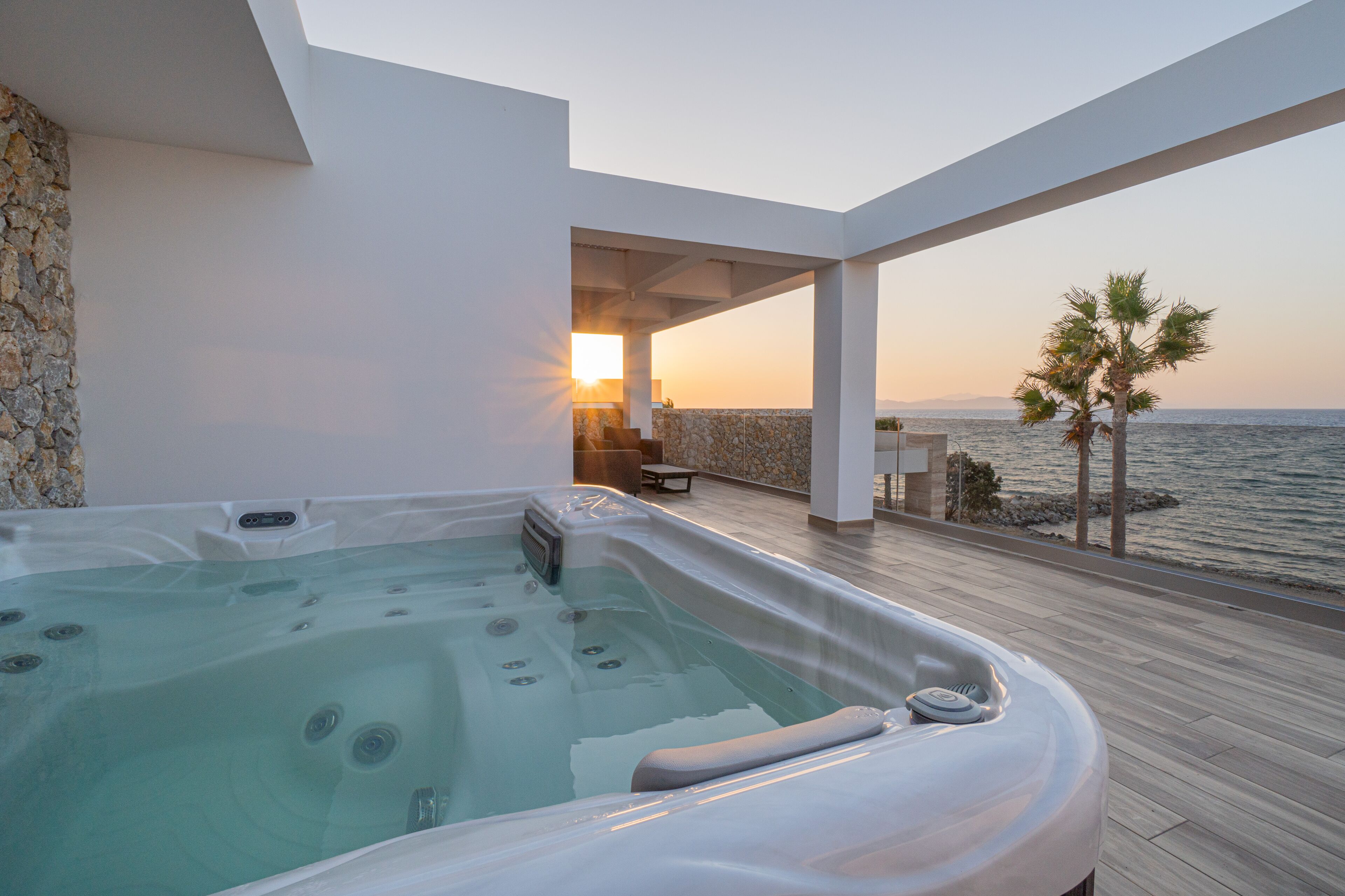 Ein moderner Wellnessbereich mit Whirlpool, Steinwand und Blick auf das Meer bei Sonnenuntergang.