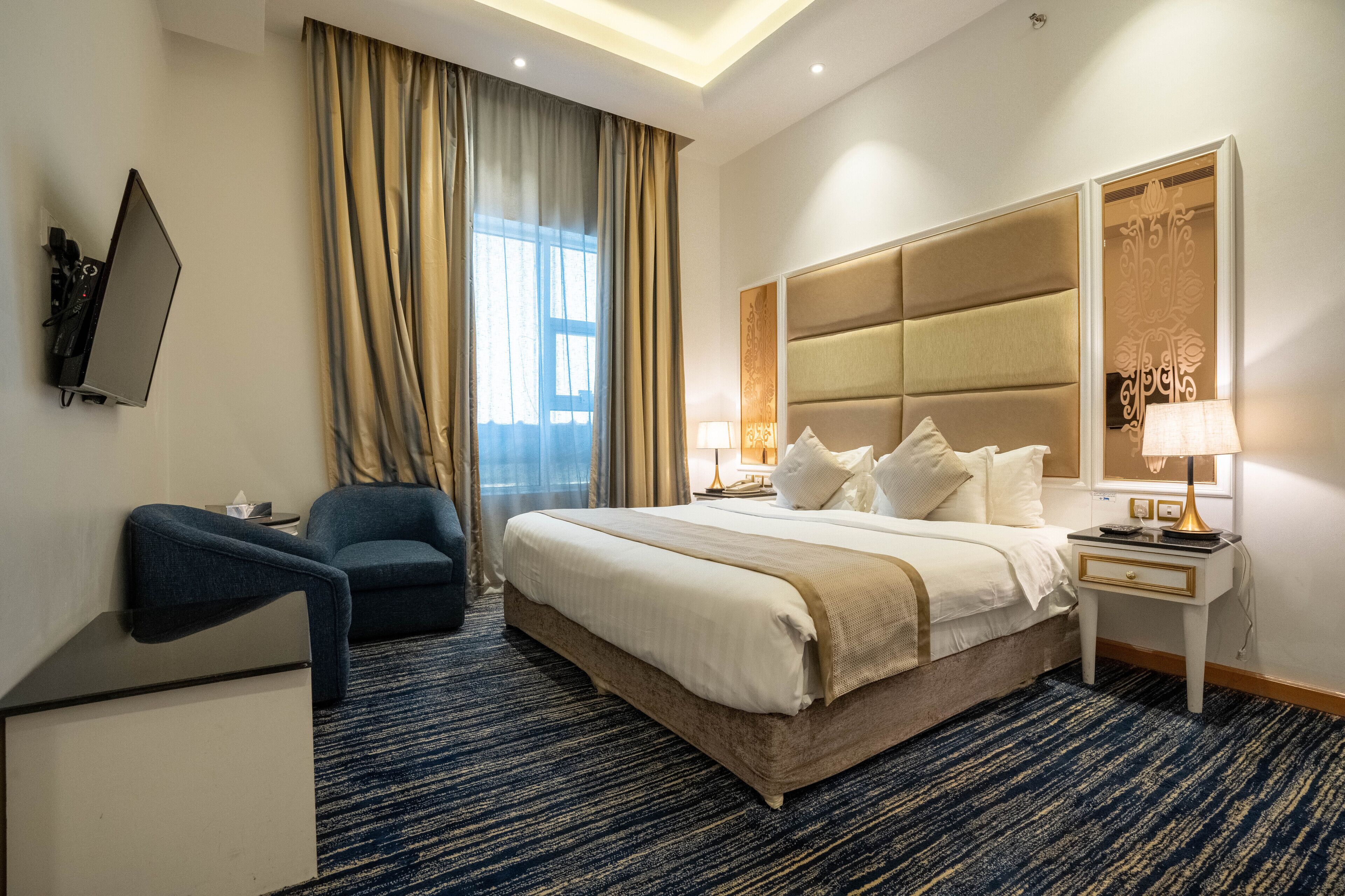 A hotel room with a large bed, a desk with a lamp, a chair, and a TV mounted on the wall.