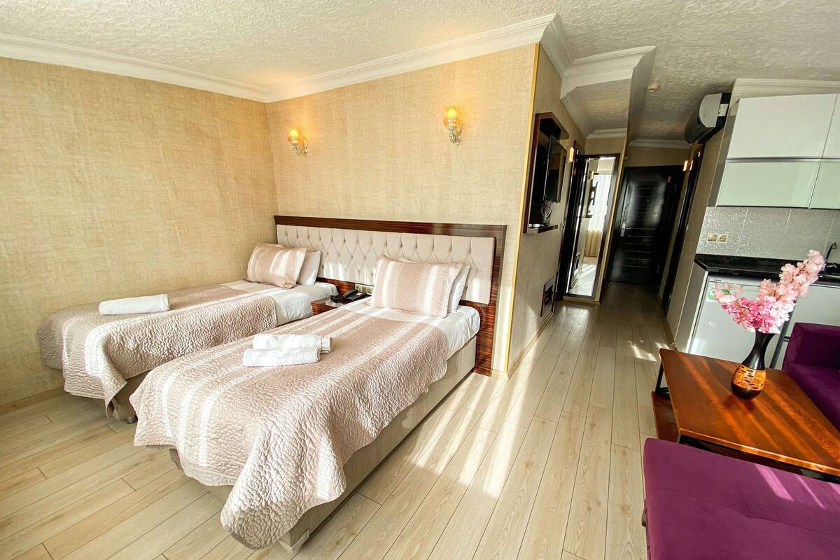 A hotel room with two beds, a wooden floor, a purple armchair, and a vase with flowers.