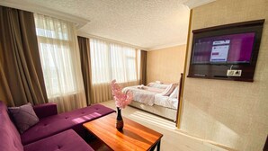 Une chambre d’hôtel avec un canapé, une table en bois, un vase de fleurs, un lit avec une couverture et un téléviseur fixé au mur.