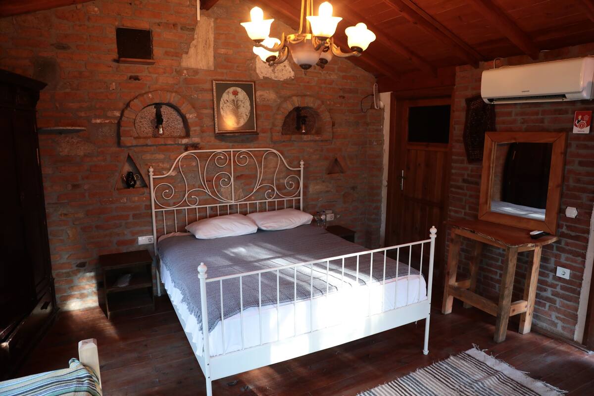 Une chambre avec un mur en briques, un cadre de lit en métal blanc, un lustre et une table en bois.