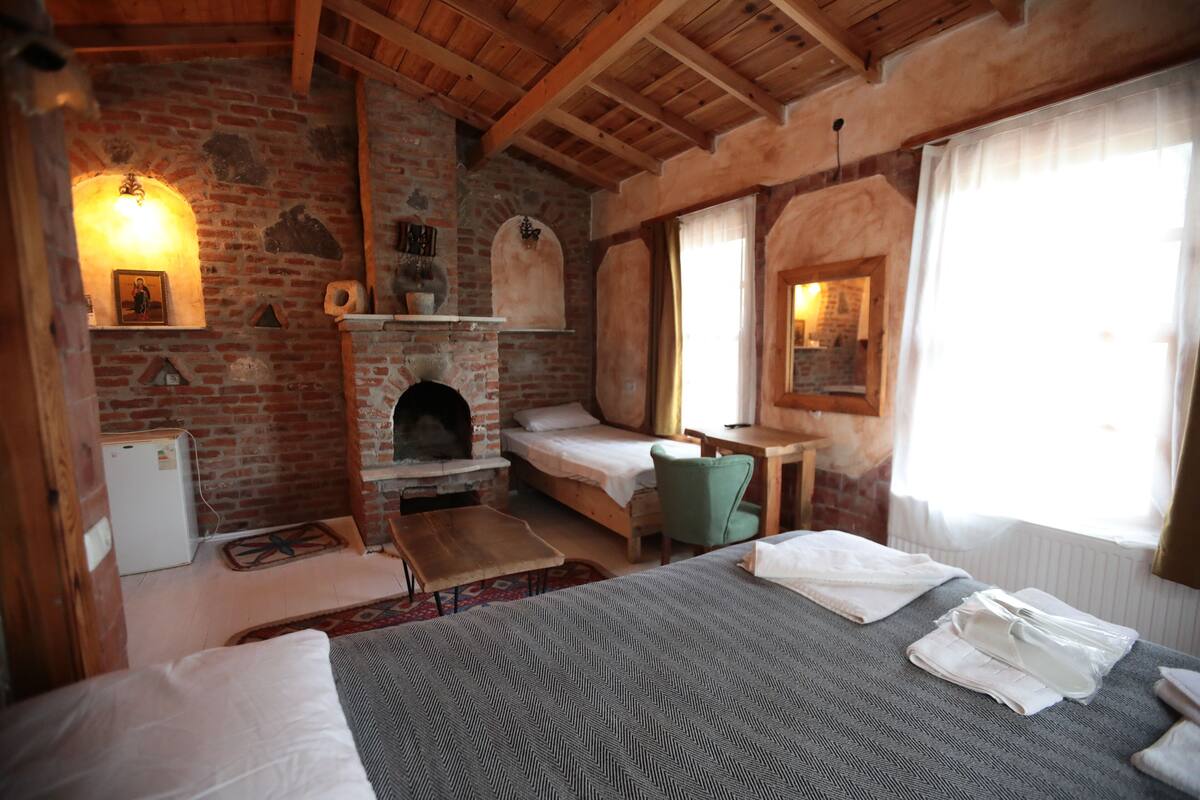 Une pièce rustique avec une cheminée en briques, un lit recouvert d’une couverture à motifs, une petite table et une chaise verte.