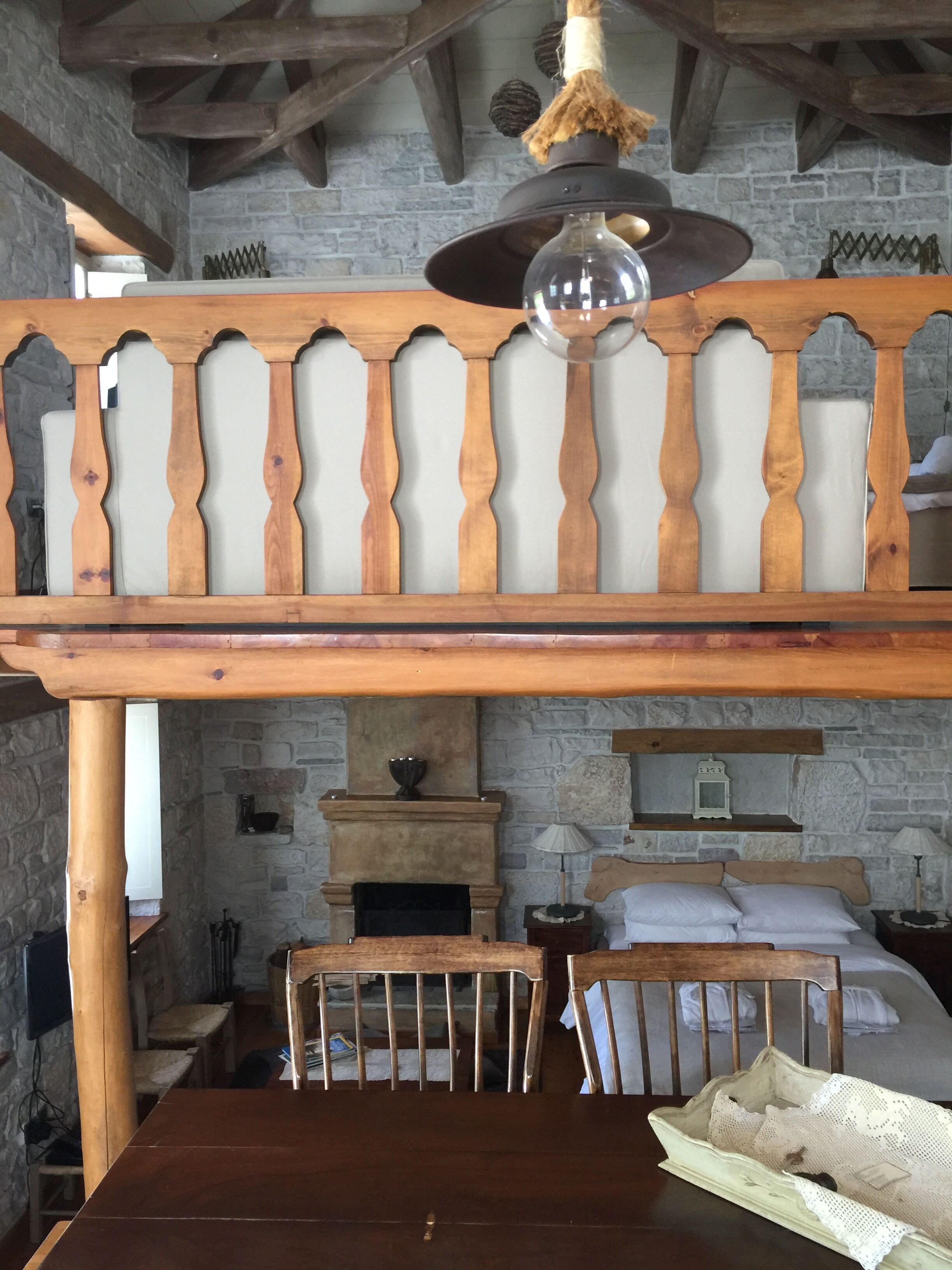 Una habitació rústica amb taula de fusta, cadires, llum penjant i una llar de pedra.