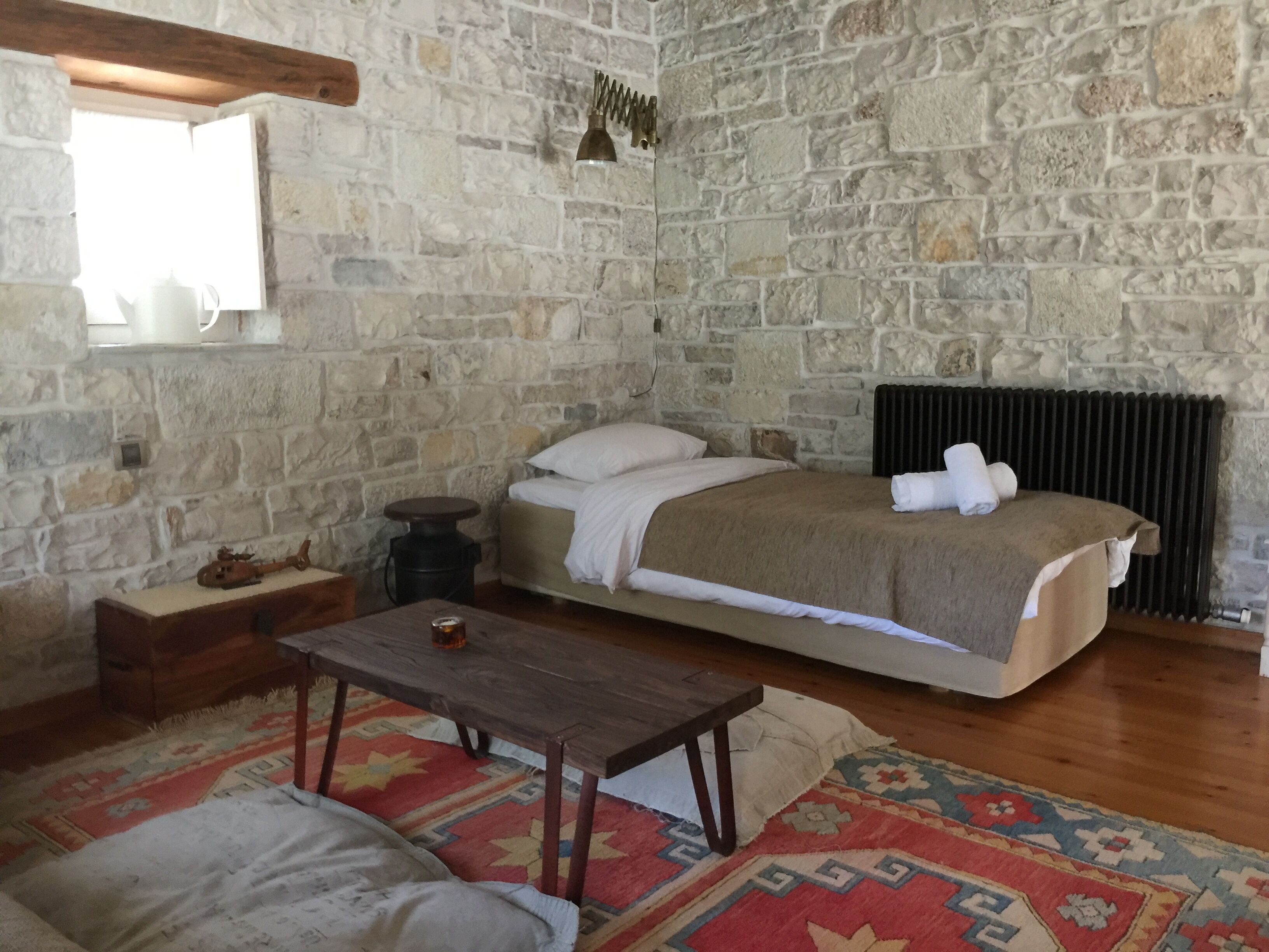 A room with a stone wall, a bed with a white blanket, a wooden table, and a patterned rug.