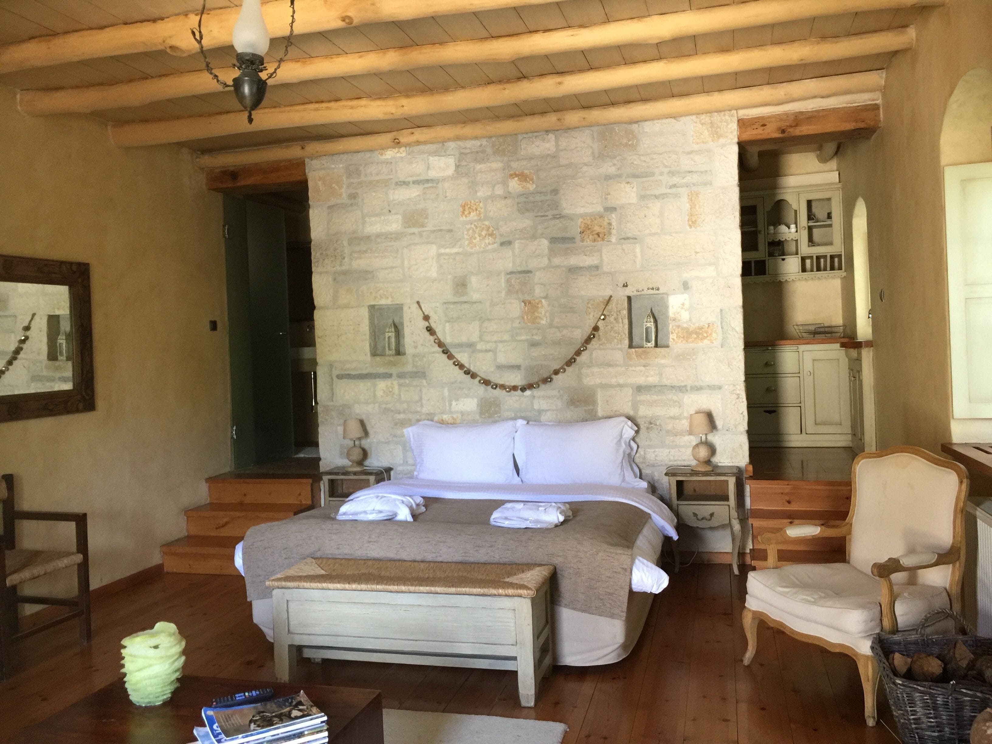 A cozy bedroom with a stone wall, wooden beams, a bed with white linens, a bench, a chair, and a small table with books.