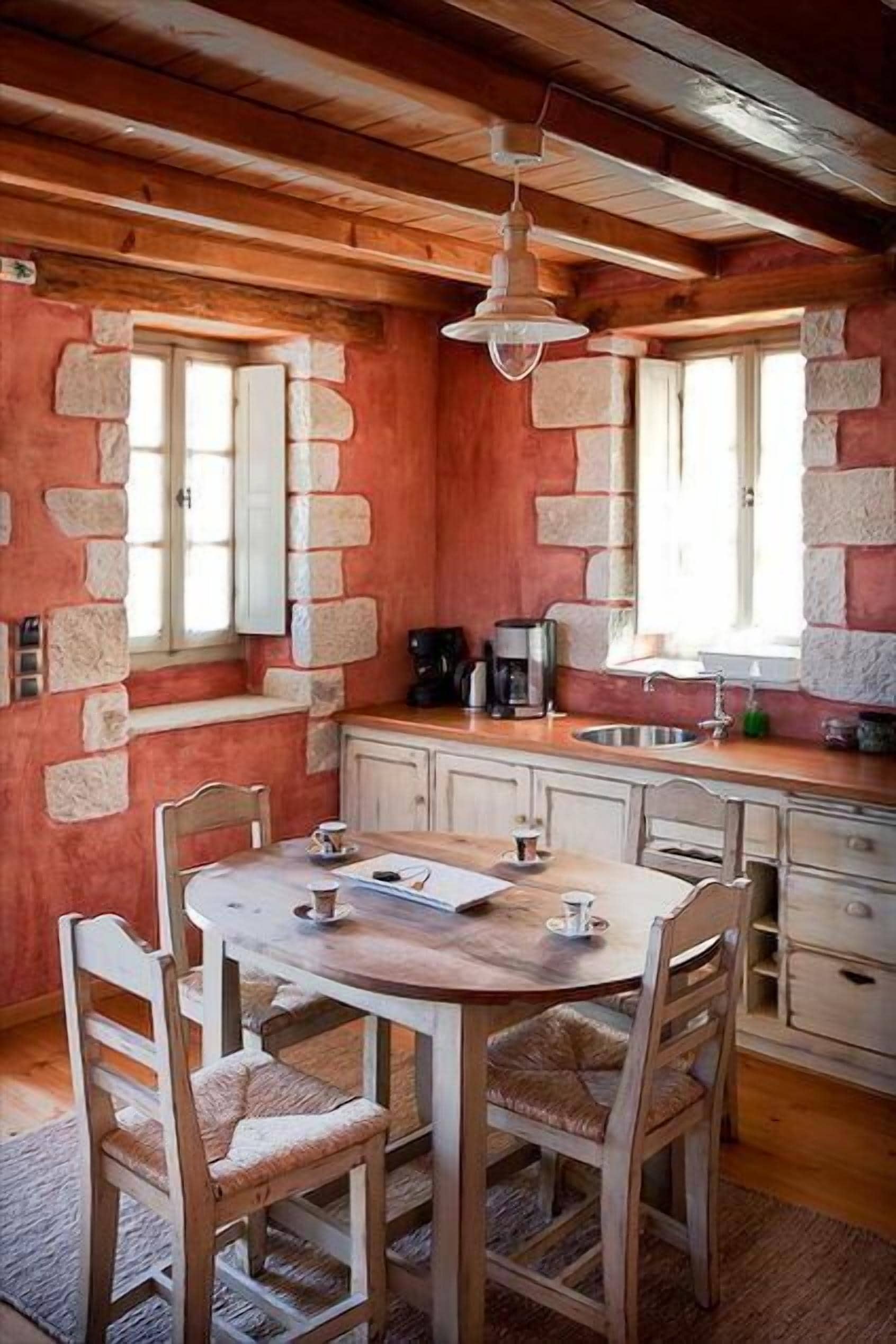 A rustic kitchen with stone walls, wooden ceiling beams, a round wooden table with chairs, a hanging light fixture, and a window with a stone frame.