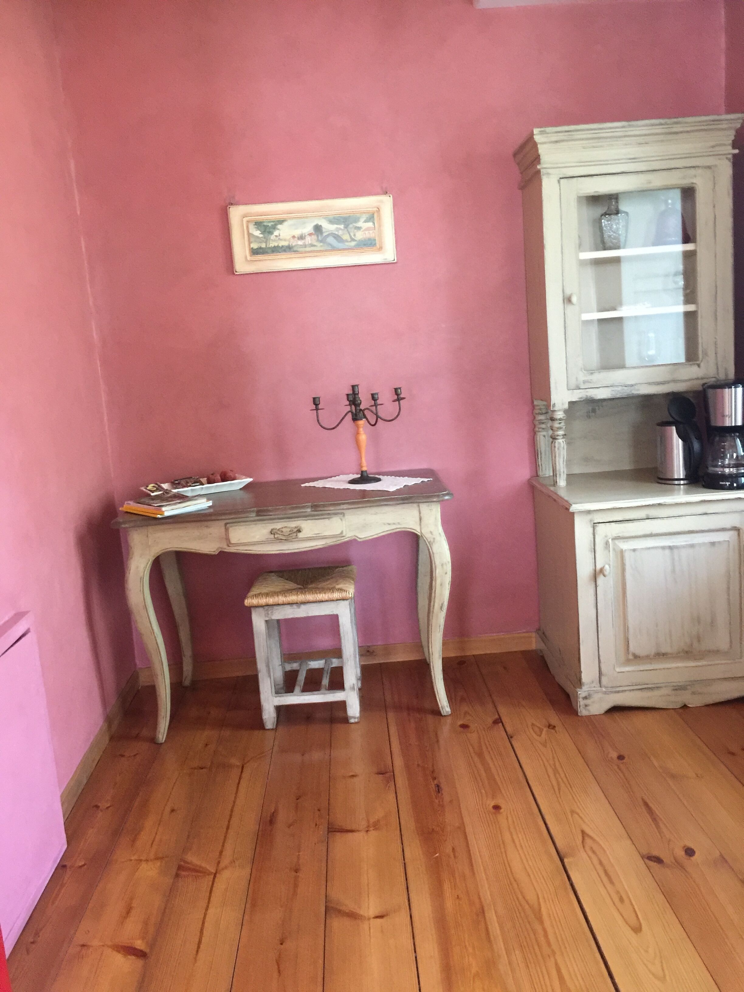 A room with a wooden floor, a vintage-style desk with a candelabra, a small stool, and a cabinet with a glass door.