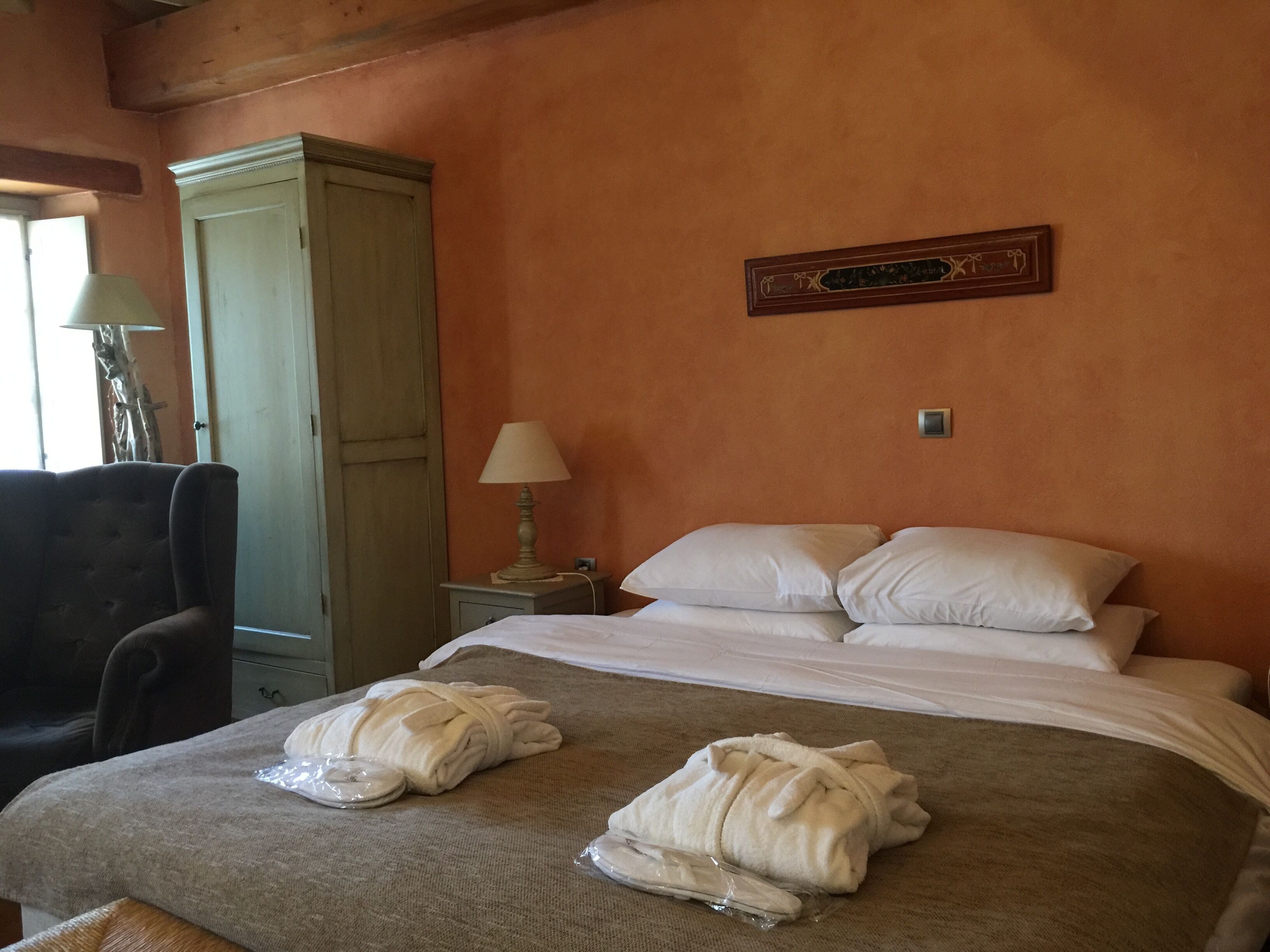 Quarto de hotel com cama, duas almofadas brancas, luminária de cabeceira, guarda-roupa de madeira e um objeto decorativo na parede.