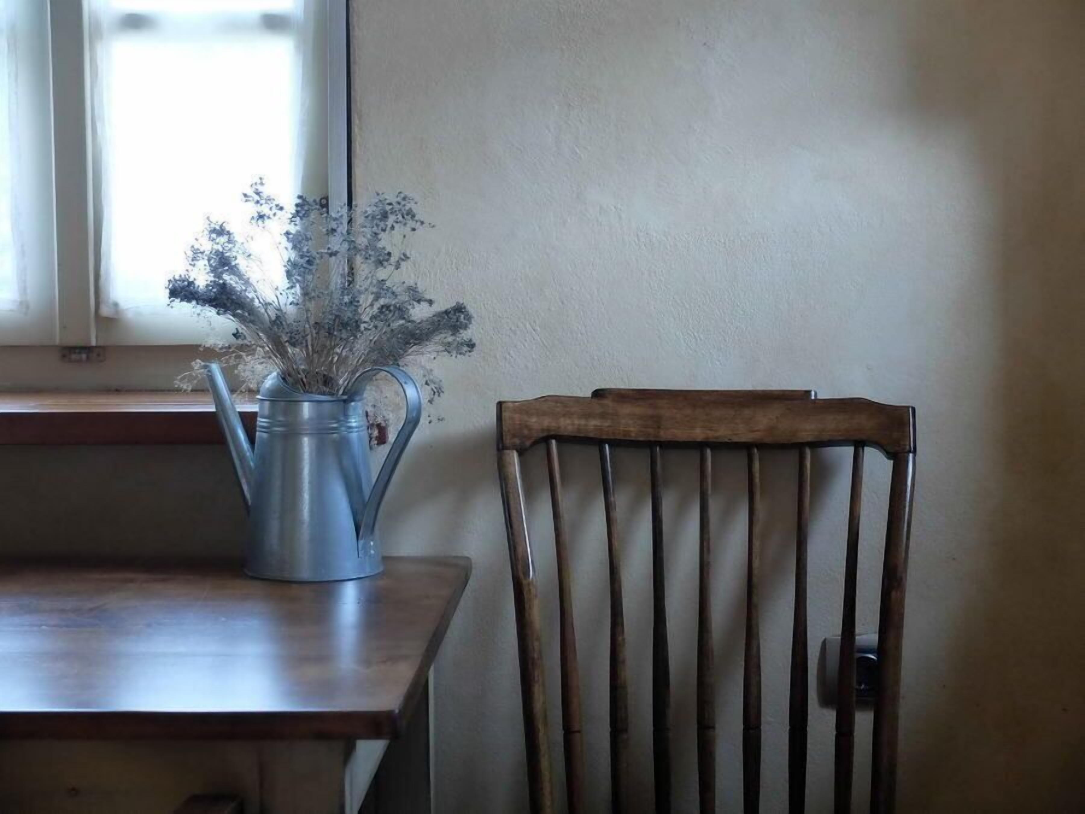 Una silla de madera con respaldo de listones, colocada junto a una mesa con una jarra de metal que contiene flores secas.