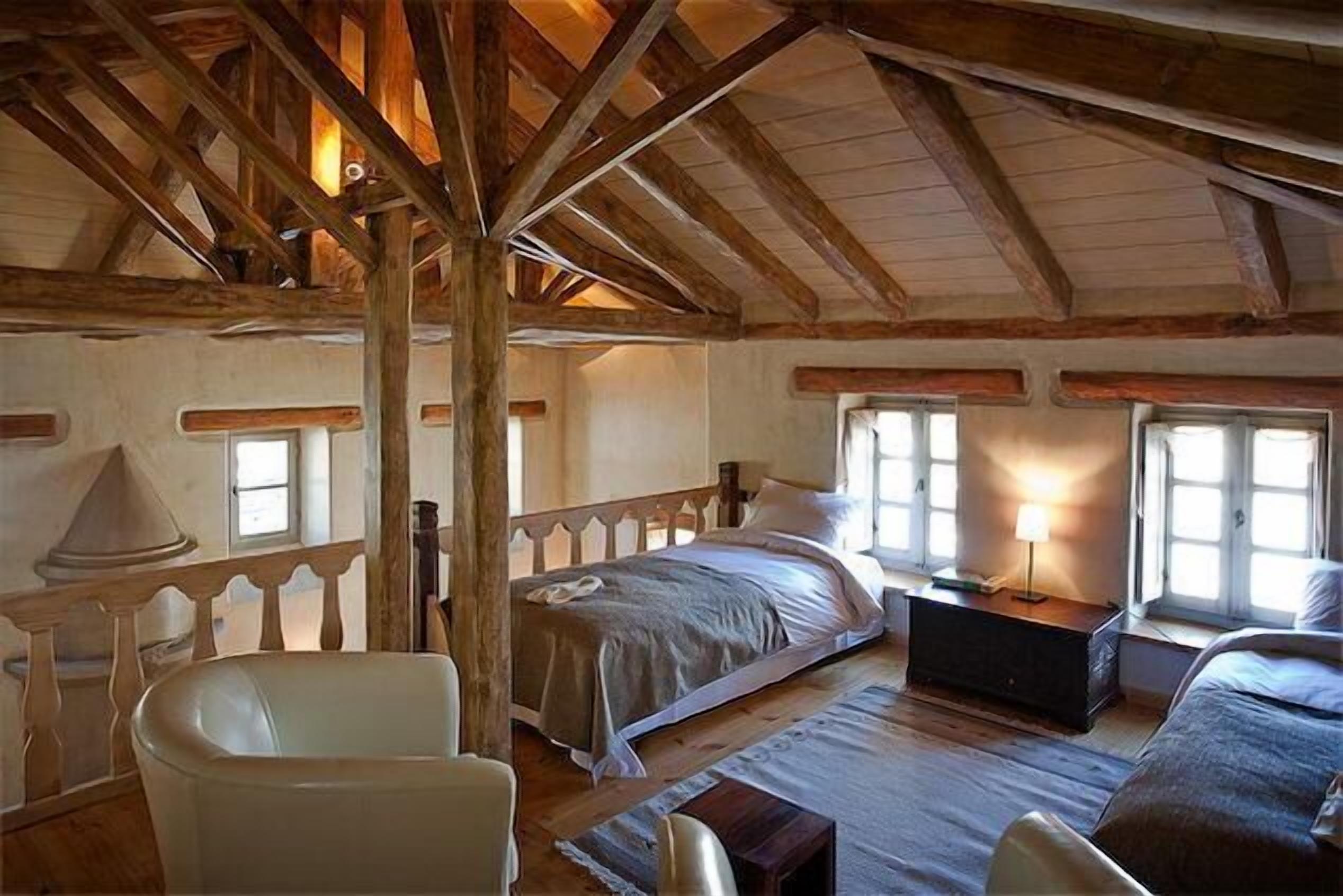 A rustic bedroom with a wooden ceiling, a bed, a nightstand, a chair, and a window with natural light.
