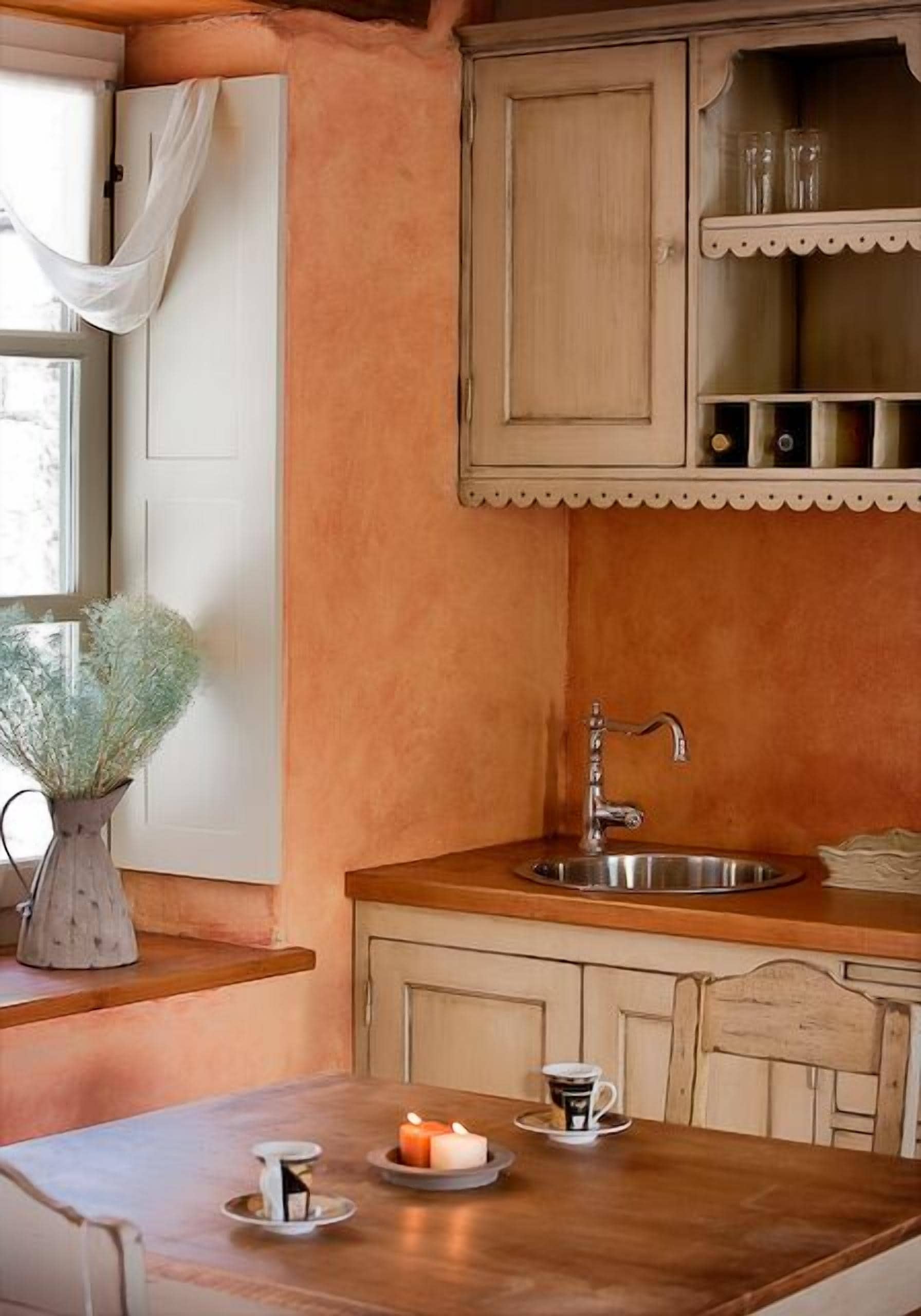 A kitchen with a wooden countertop, a sink, and cabinets. There is a window with a white curtain, a vase with dried flowers, and a candle on the table.