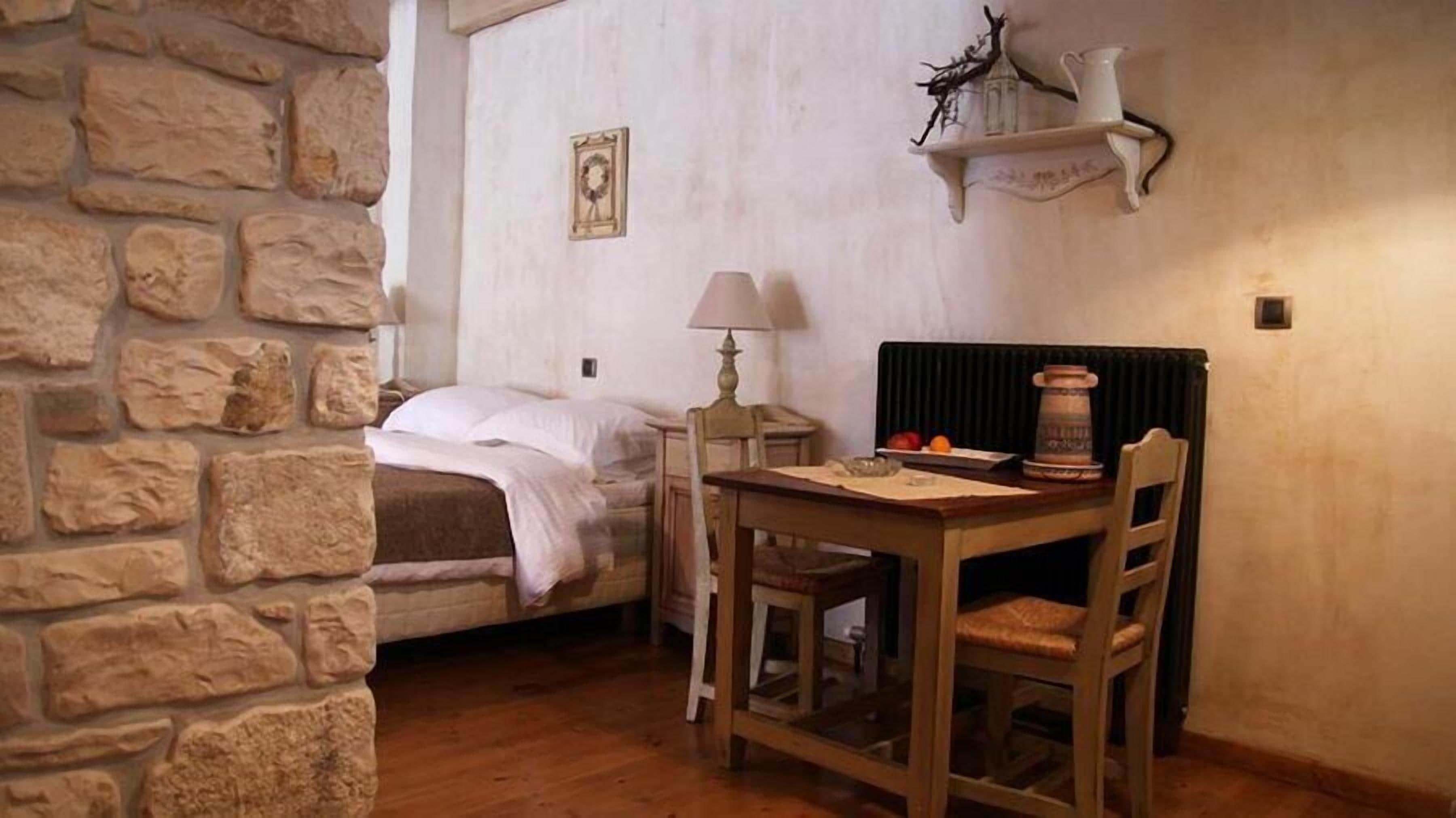 Una habitación acogedora con pared de piedra, cama, una mesa con sillas, lámpara y estante con adornos.